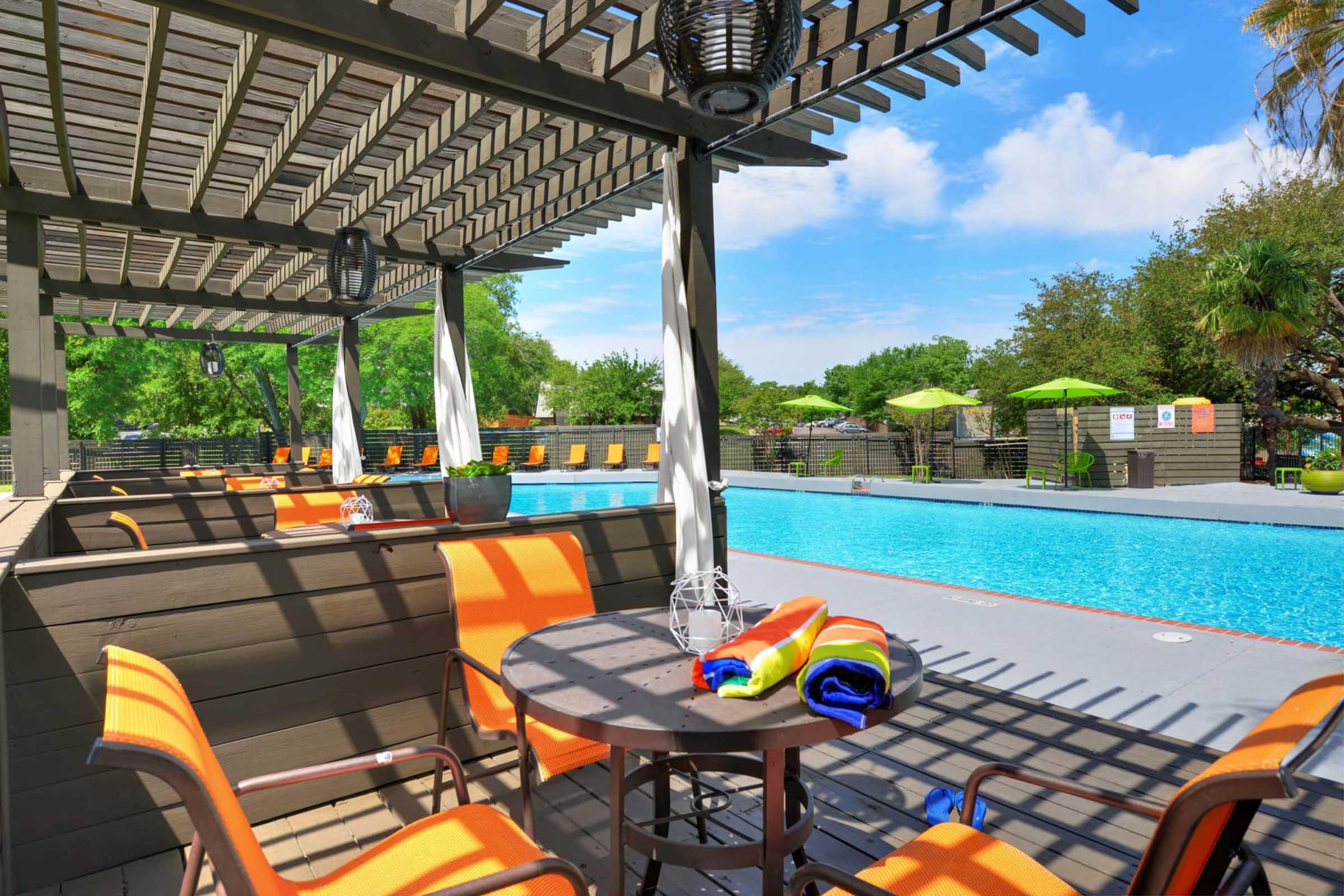 A sunny poolside scene featuring a sparkling blue pool surrounded by lounge chairs with orange cushions. There are umbrellas providing shade, and colorful towels are neatly arranged on a table. Lush greenery and a clear blue sky enhance the relaxing atmosphere of this outdoor space.