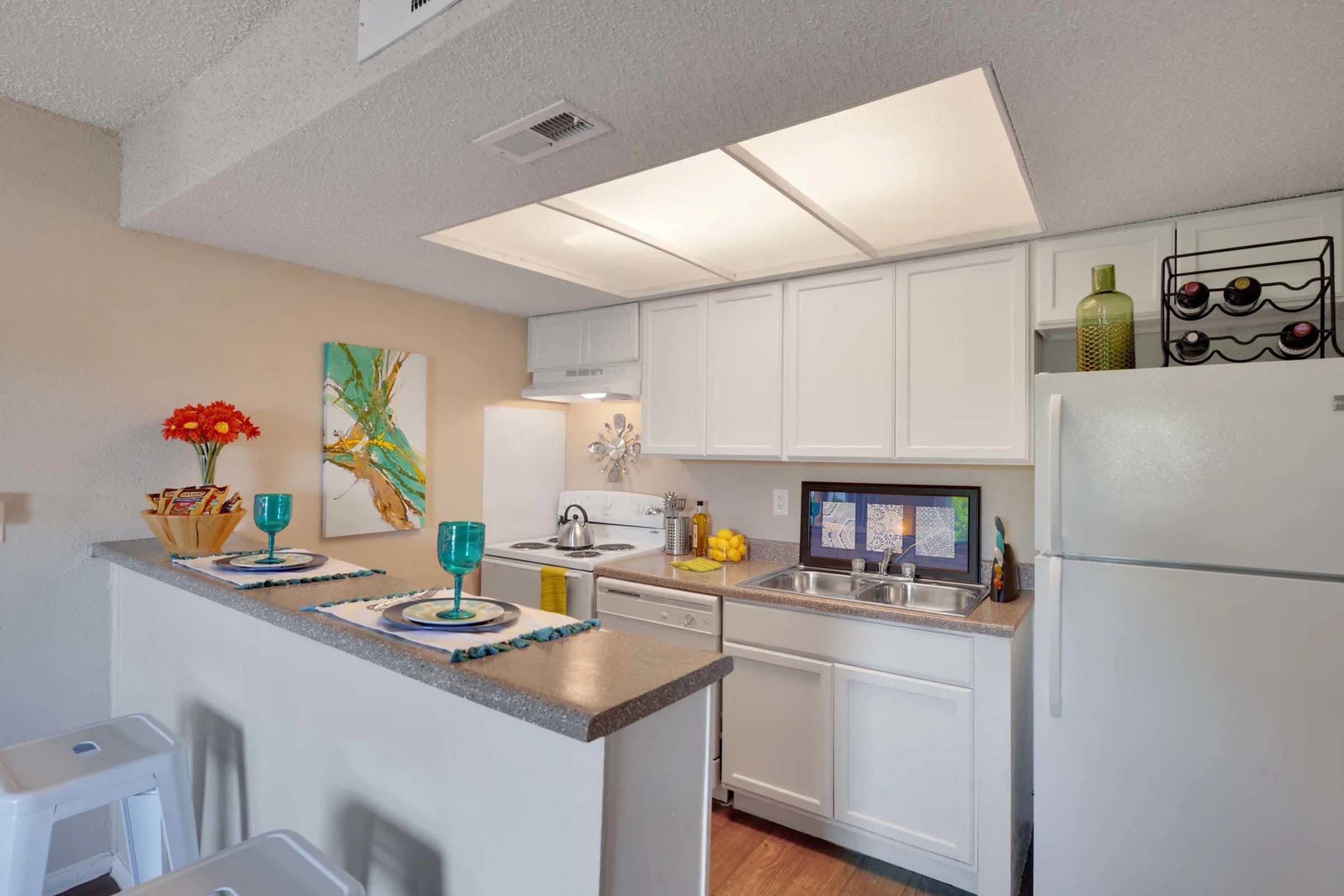 Modern kitchen featuring white cabinetry, stainless steel sink, and a refrigerator. The countertop holds turquoise glasses, a lemon centerpiece, and decorative items. A painting with vibrant colors adorns the wall. The space is brightly lit, creating a welcoming atmosphere.