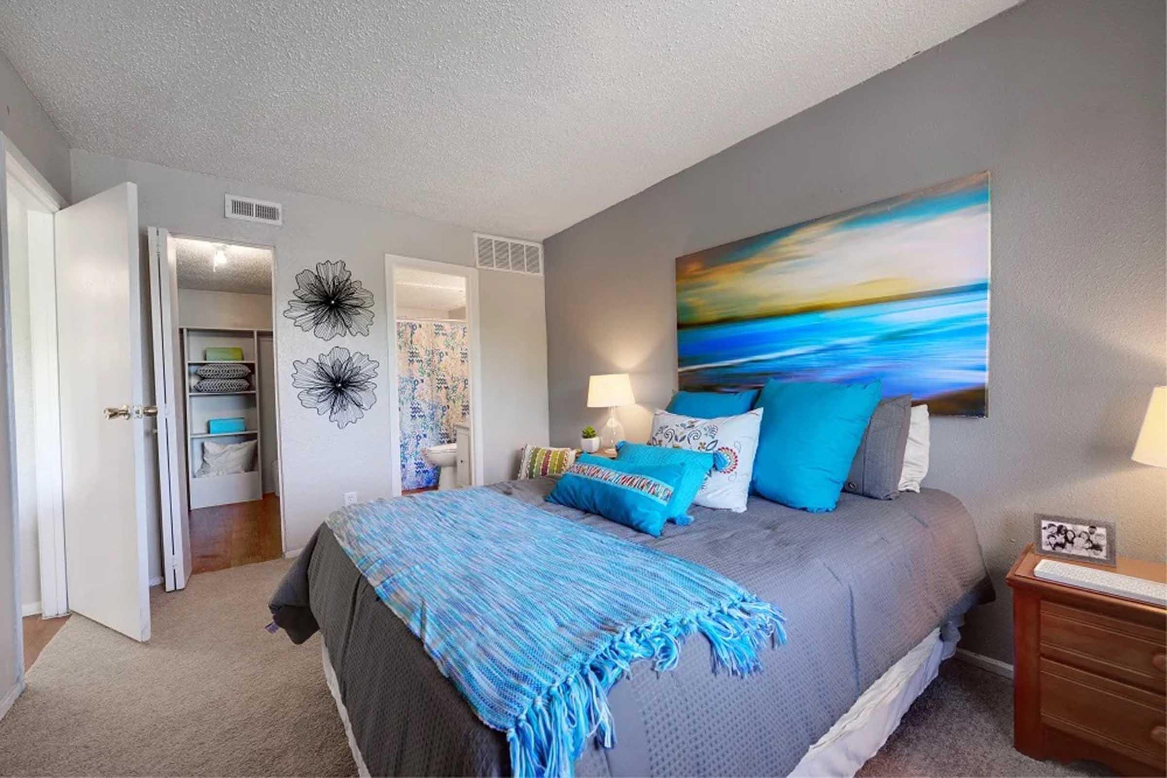 A cozy bedroom featuring a large bed with blue and gray bedding, accented by colorful pillows. The room has a nightstand with a lamp and a decorative photo. The walls are painted a soft gray, and a colorful abstract painting hangs above the bed. A bright bathroom is partially visible through an open door.