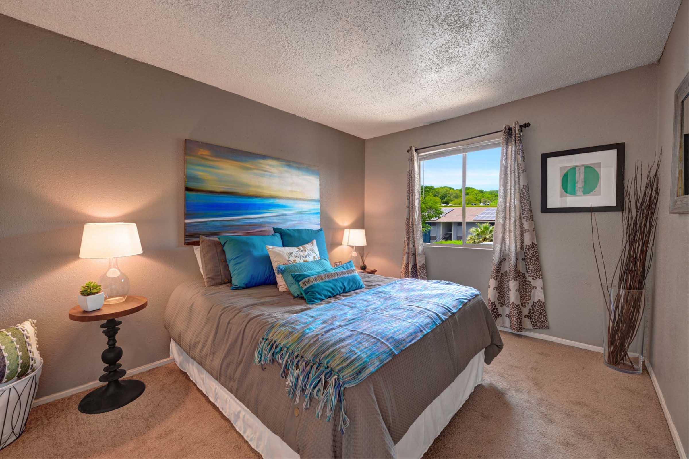 Cozy bedroom featuring a queen-sized bed with blue and teal decorative pillows, a colorful abstract painting above the bed, and bedside lamps. A large window lets in natural light, and a decorative plant sits on a nightstand. The walls are painted in a neutral tone, enhancing a warm and inviting atmosphere.