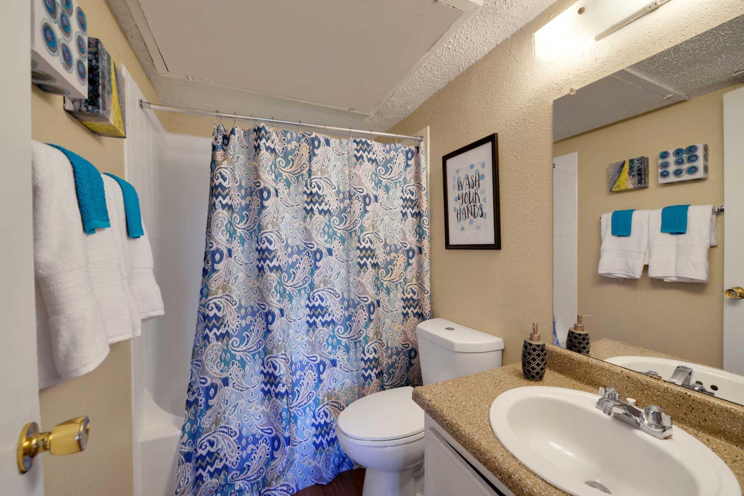 A small bathroom featuring a patterned shower curtain in blue and white, a toilet, and a stone countertop with a sink. Two sets of white towels are hanging on the wall, and there are decorative items on the counter. The walls are painted a light color, and there is artwork and decorative accents mounted on the walls.