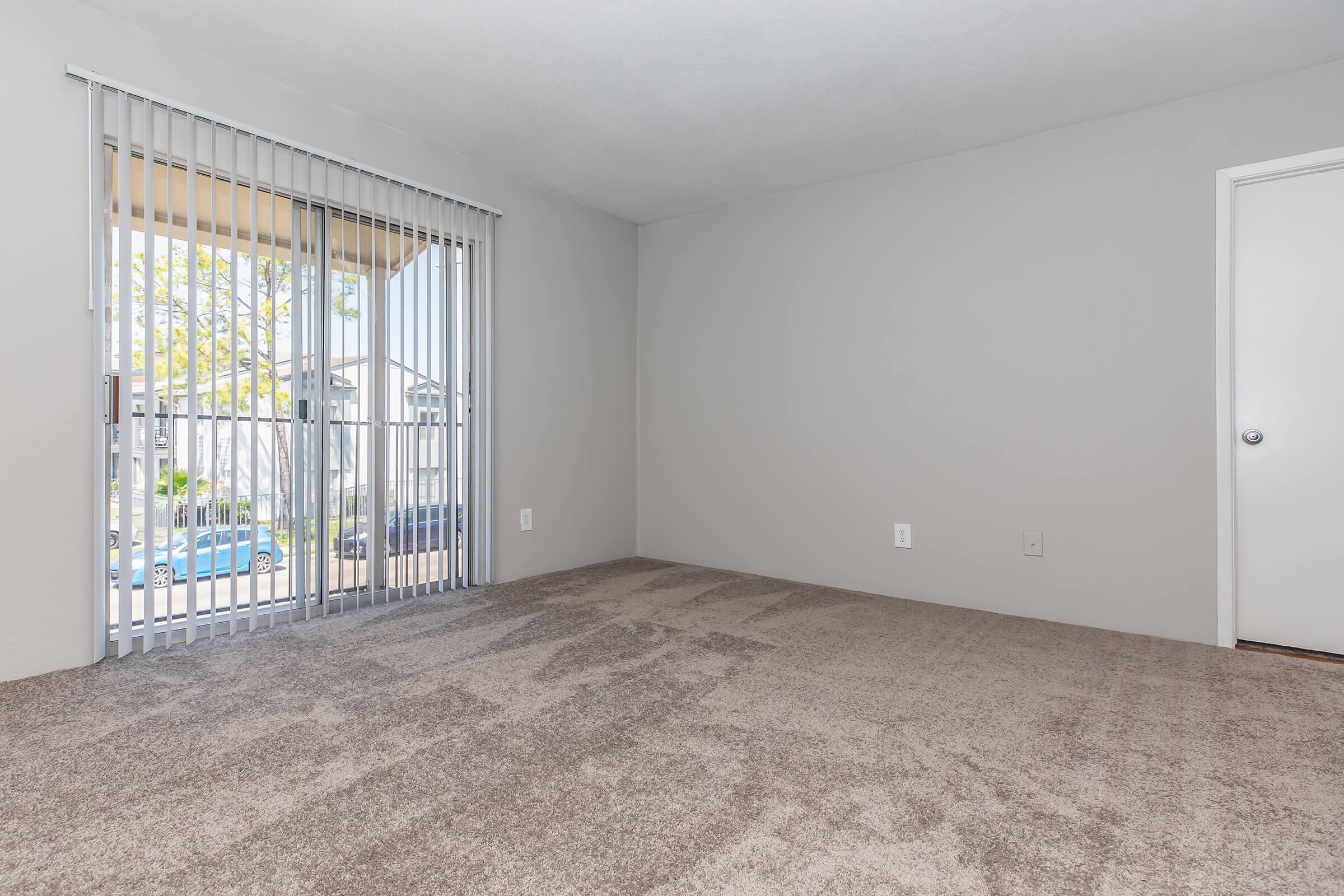 A spacious, empty room with light gray walls and soft, beige carpet. There are large sliding glass doors covered with vertical blinds, leading to a balcony. A doorway on the right suggests access to another room, and natural light brightens the space, creating an inviting atmosphere.