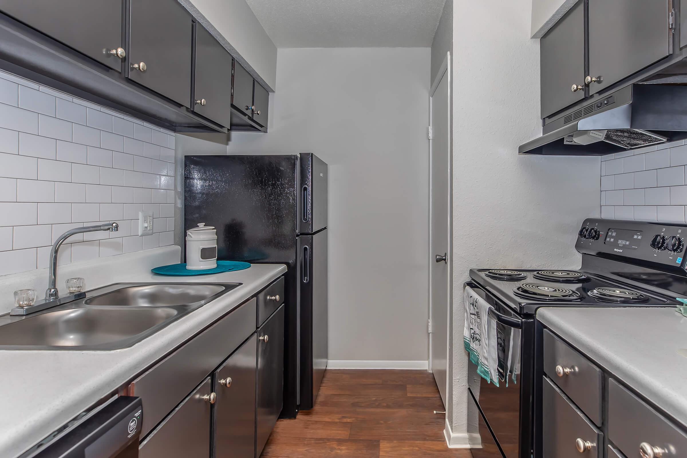 A modern kitchen featuring dark cabinetry, a double sink with a faucet, a black refrigerator, and a black stove with four burners. The kitchen has white tiled backsplashes and wooden flooring, creating a clean and contemporary look. A door is visible at the end of the kitchen space.