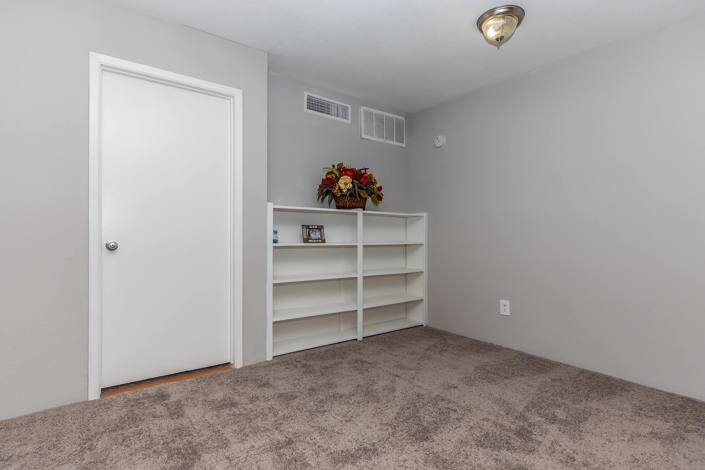 A neutral-toned room with gray walls and carpet. There's a white bookshelf against the wall, decorated with a small photo frame and a flower arrangement on top. A closed door is visible on the left side of the image, and a ceiling light fixture is installed above.