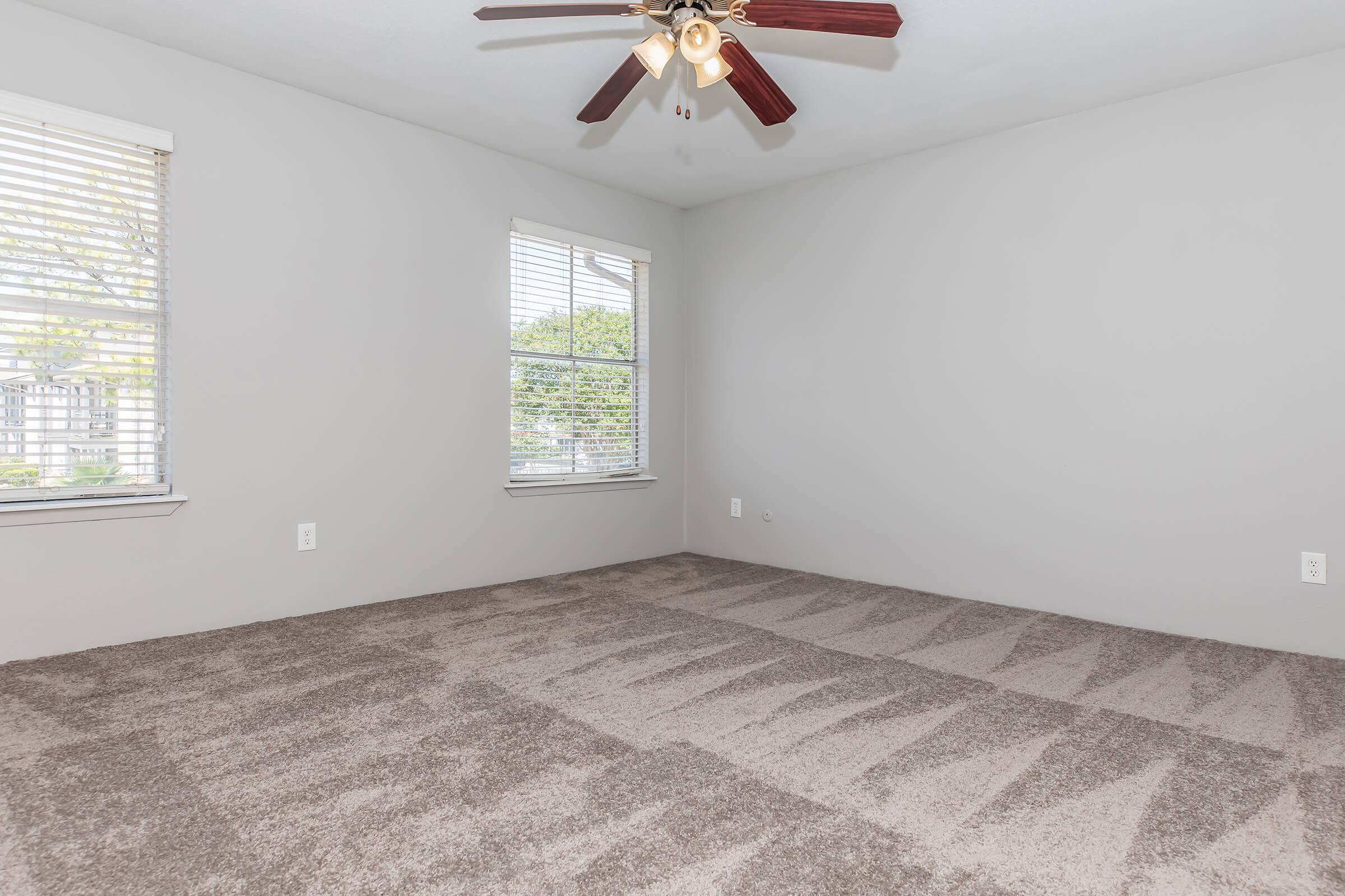 A spacious, empty room with light gray walls and large windows, allowing natural light to enter. The floor is covered with plush, light-colored carpet, and a ceiling fan with wooden blades is mounted above. The room is unfurnished, creating a bright and open atmosphere.