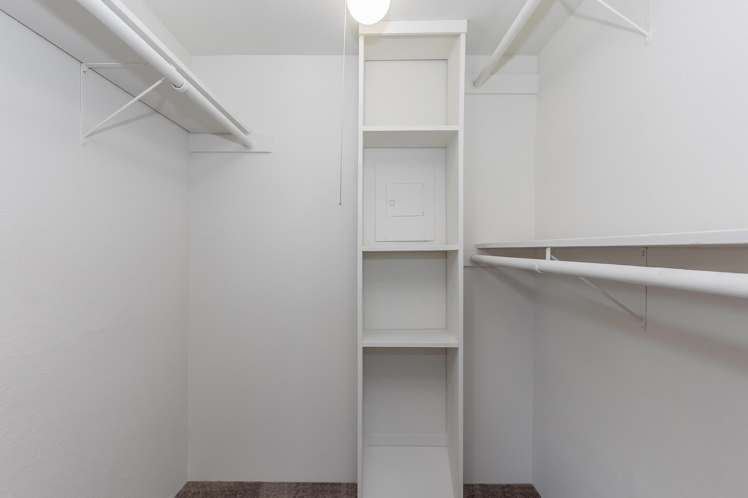 A minimalistic empty closet with white walls and shelving. The closet features two hanging rods on either side and a tall shelving unit in the center, with an overhead light illuminating the space. The floor is carpeted in a neutral tone, enhancing the simplicity of the design.