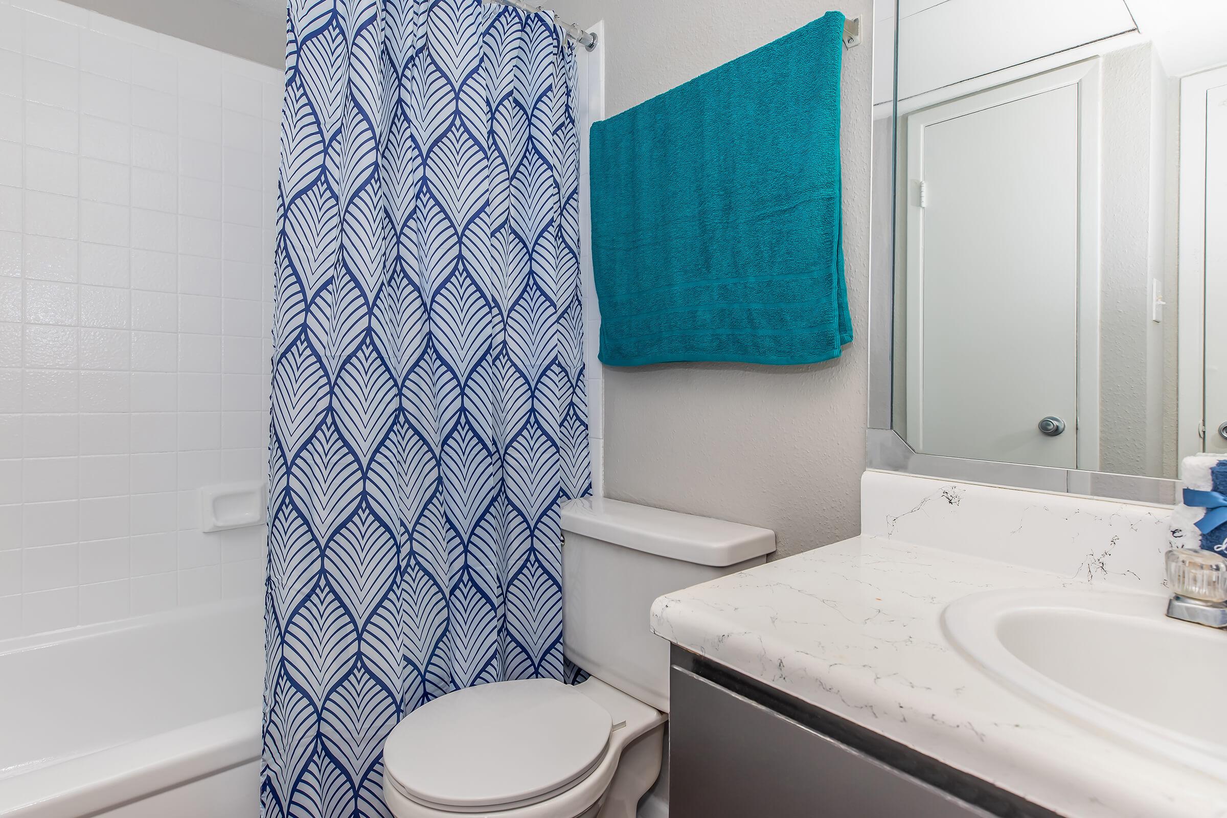 A modern bathroom featuring a shower with a blue and white leaf-patterned curtain, a white toilet, and a marble countertop with a sink. A bright teal towel hangs on the wall, and there is a large mirror reflecting part of the room. The walls are painted in a neutral tone, enhancing the clean and fresh ambiance.