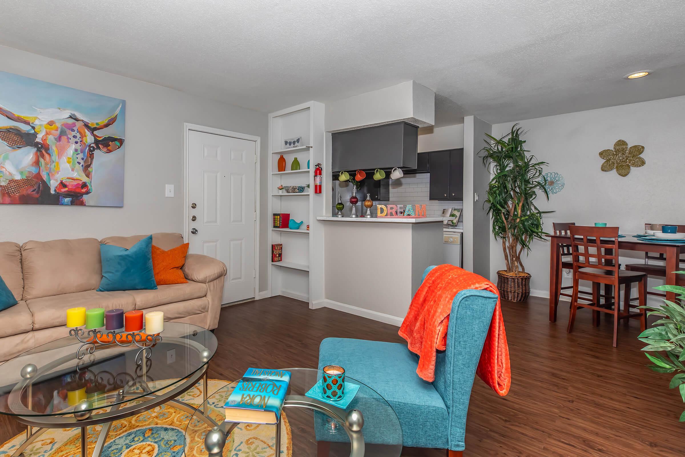 A cozy living room featuring a beige sofa with colorful cushions, a glass coffee table, and a vibrant painting of a cow on the wall. The kitchenette is open to the living area, with modern appliances and colorful decor. A potted plant and dining table with chairs complete the inviting space.