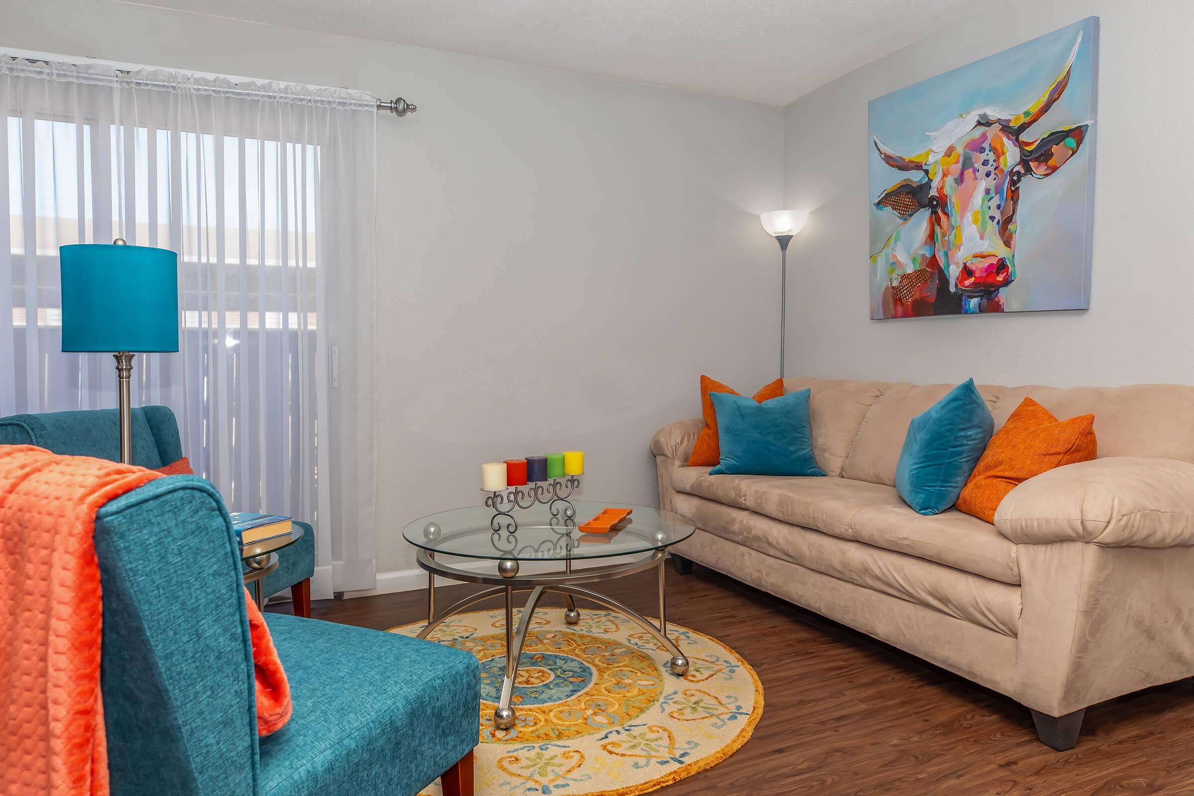A cozy living room featuring a beige sofa with colorful cushions, a glass coffee table, and vibrant decor. There are two teal armchairs, a turquoise lamp, and a circular patterned rug. A large painting of a colorful cow hangs on the wall, enhancing the room's cheerful atmosphere. Natural light filters through sheer curtains.