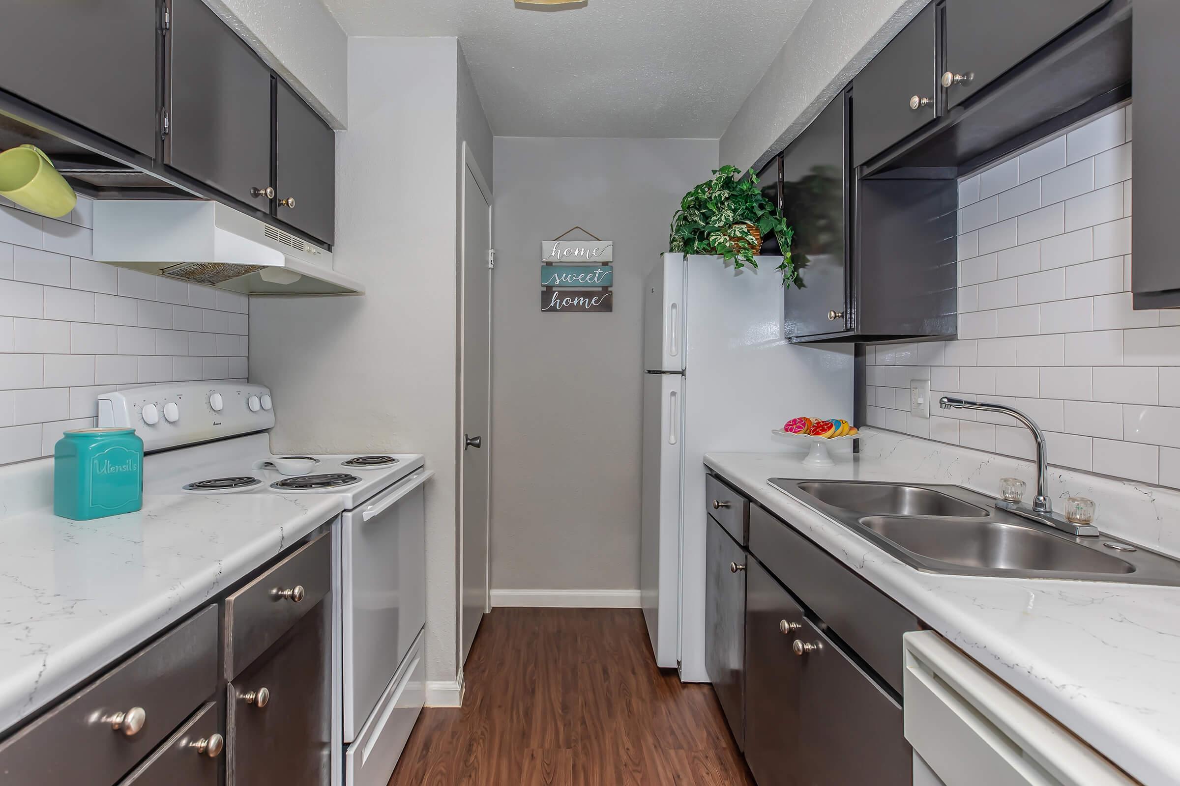 A modern kitchen featuring dark cabinetry, white countertops, and stainless steel appliances. The space includes an electric stove, oven, and a double sink. A refrigerator and decorative wall art add to the inviting atmosphere, complemented by a small potted plant and colorful kitchen accessories.