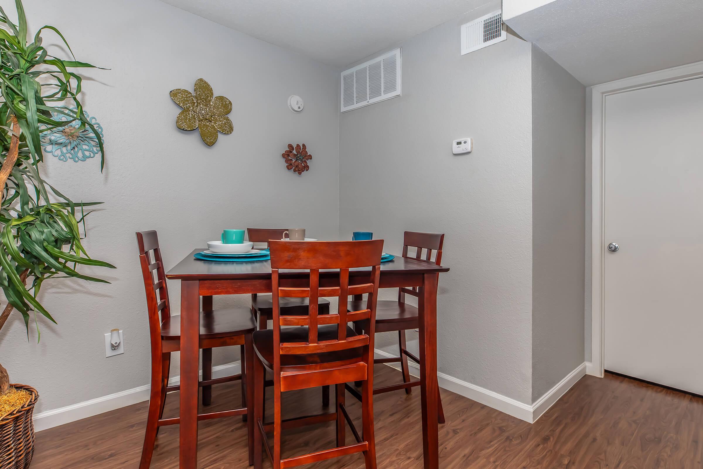 A cozy dining area featuring a wooden table with four high-backed chairs, set with blue plates and utensils. The walls are painted a light gray, adorned with decorative flowers, and a green potted plant adds a touch of color. A thermostat on the wall and a closed door complete the scene.
