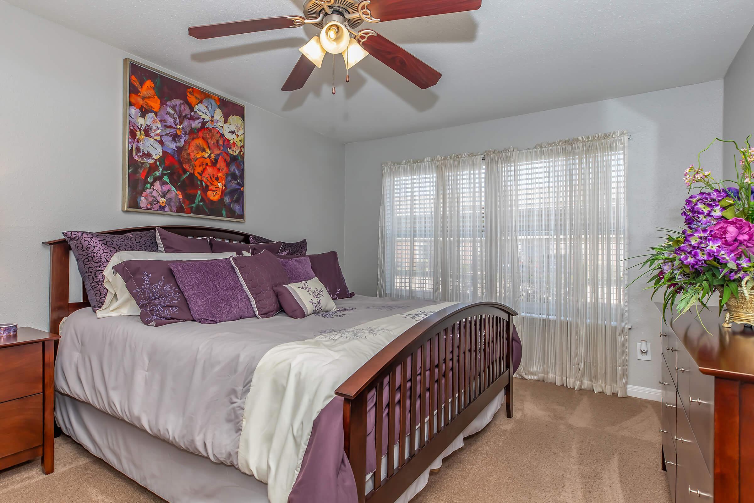 A cozy bedroom featuring a large wooden bed with purple and gray bedding, complemented by decorative pillows. A colorful flower painting hangs on the wall, while a ceiling fan adds a stylish touch. The room has a large window with sheer curtains, allowing natural light to fill the space, and a dresser with a floral arrangement.