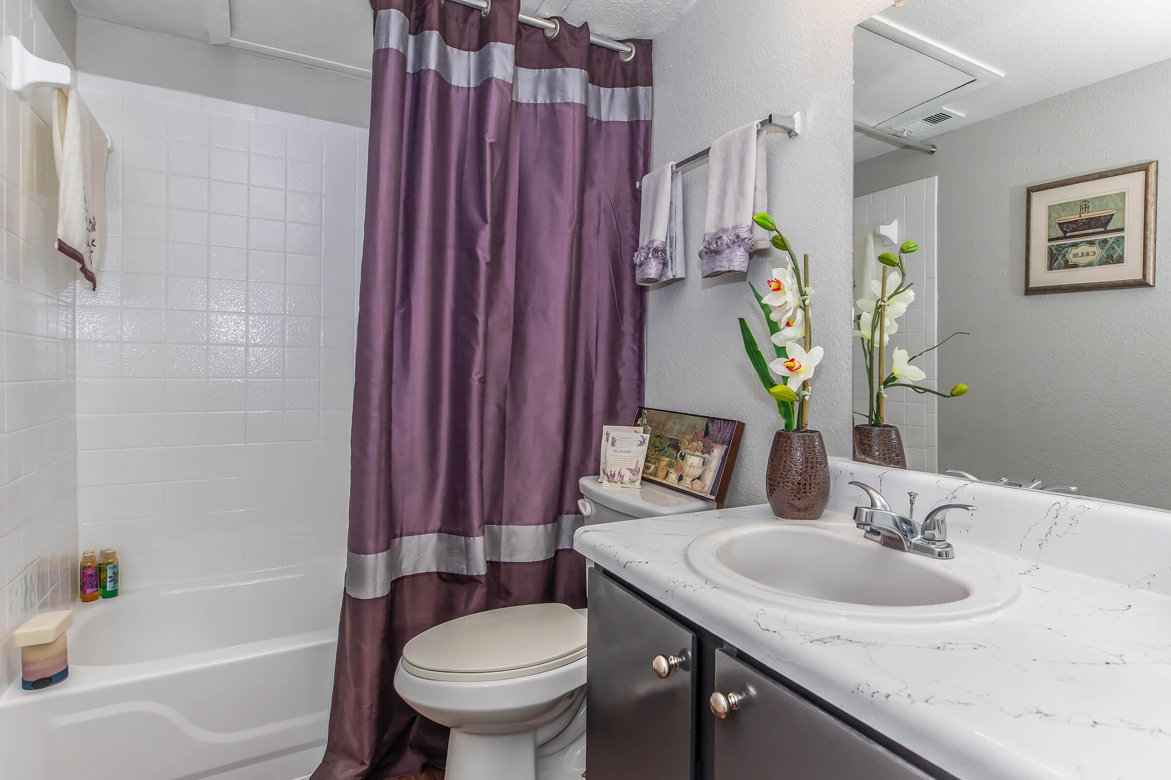 A well-decorated bathroom featuring a white bathtub with a shower curtain in shades of purple, a white countertop with a sink, and a vase with artificial flowers. There are hand towels on the wall and a framed picture on the counter, creating a clean and inviting atmosphere.