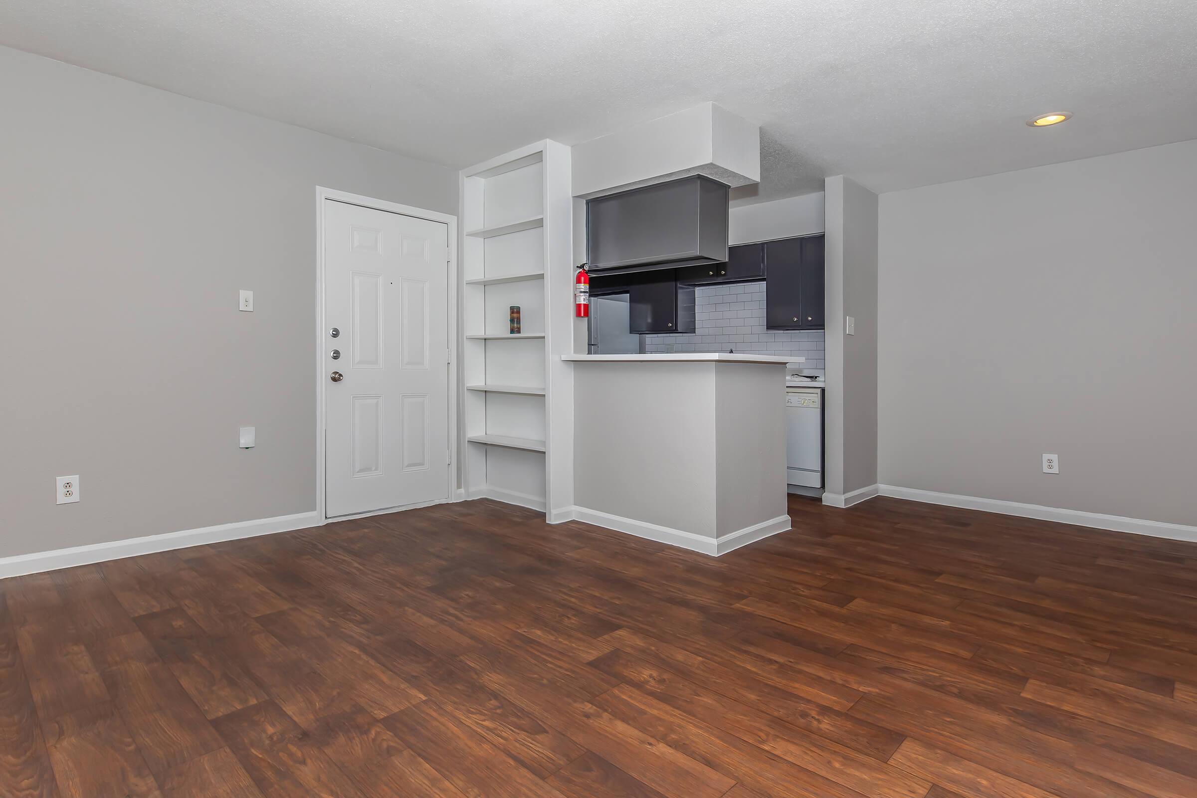 Interior view of a modern apartment featuring an open layout with hardwood flooring. A white door leads to the entrance, and a kitchen area with dark cabinetry is visible in the background. There are built-in shelves next to the kitchen, and the walls are painted a light gray. Good natural lighting enhances the space.