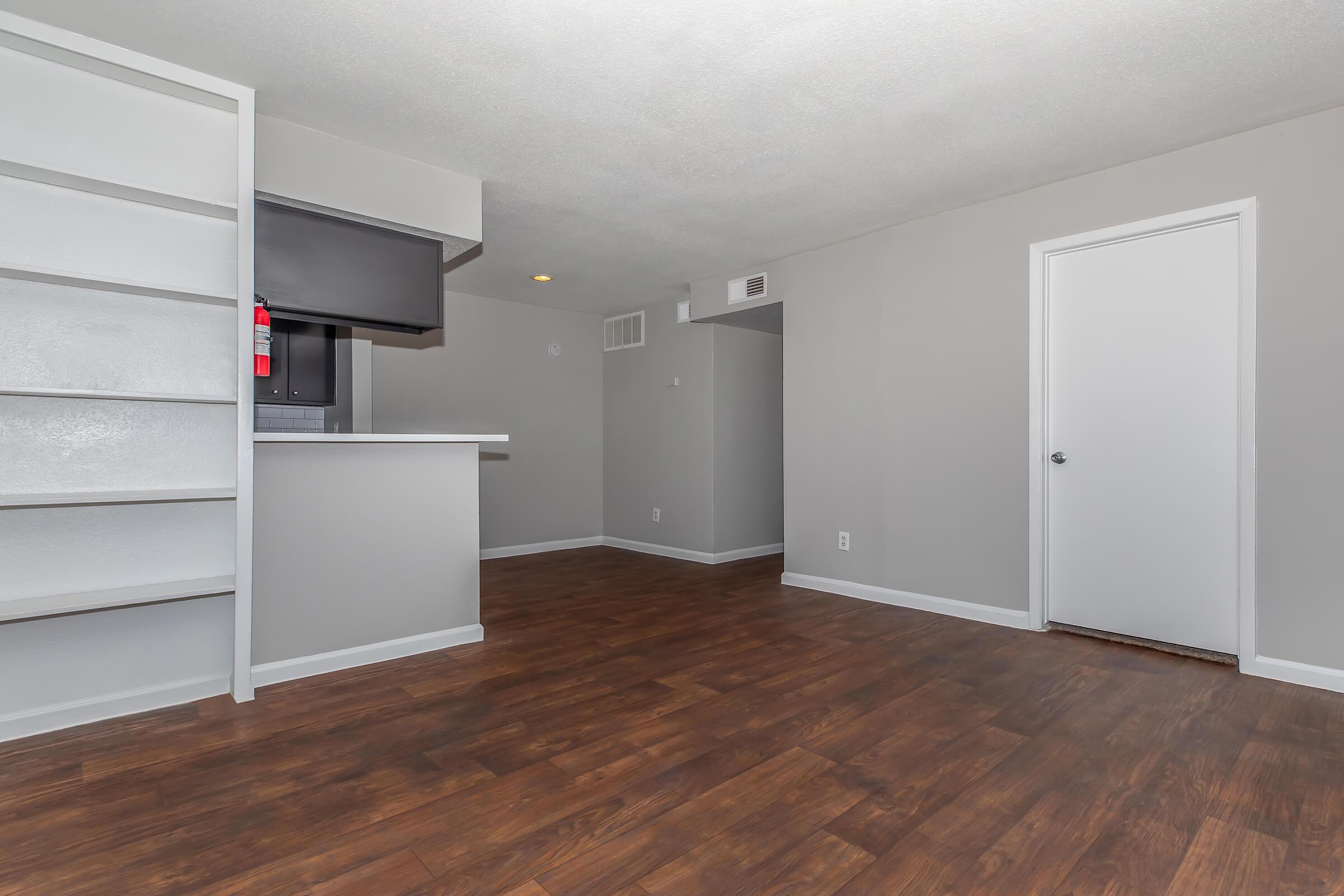 A spacious, modern living area featuring light gray walls and a wood-style floor. There is an open kitchen with a small bar area. A white door leads to another room, while a shelf unit is visible on one side. The overall ambiance is bright and inviting, suitable for comfort and relaxation.