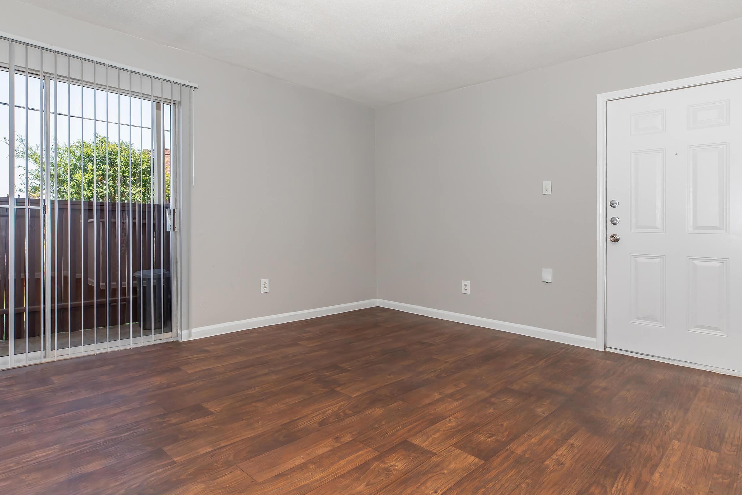Empty room with light gray walls and a hardwood floor. A sliding glass door leads to a small balcony, allowing natural light to enter. The door to the room is white with a simple design. The overall space appears clean and ready for furniture.