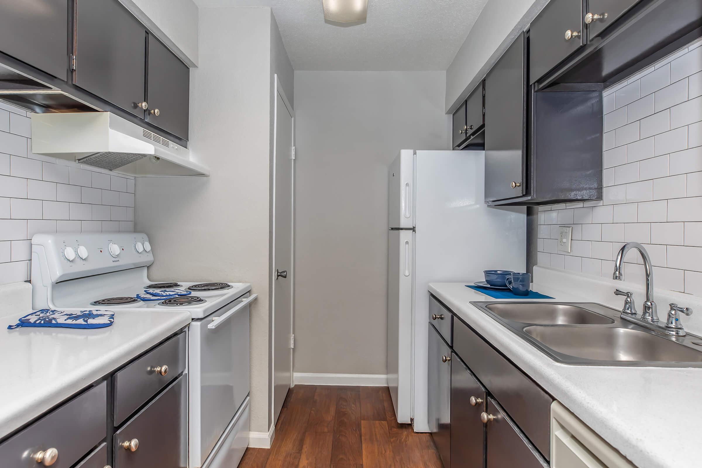 A modern kitchen with dark cabinets, white countertops, and stainless steel appliances. The layout features a stove, a refrigerator, and a sink, with tile backsplash and wood flooring. Blue dishes are placed on the counter, adding a pop of color. The space is well-lit with a light fixture above.