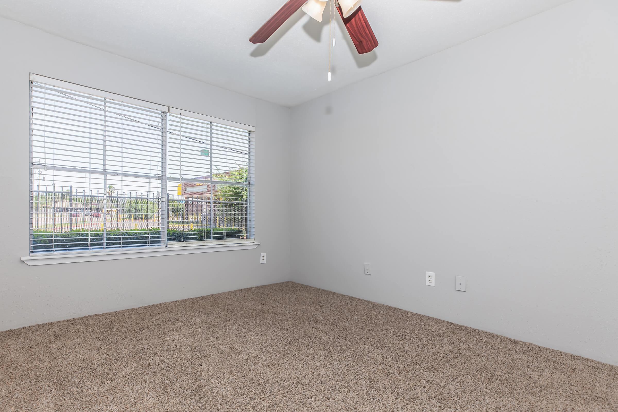 A small, empty bedroom featuring light gray walls and a tan carpet. A ceiling fan with wooden blades hangs from the ceiling. There is a window with white blinds, allowing natural light to enter, and a view of the outdoors. The space appears clean and ready for furnishings.
