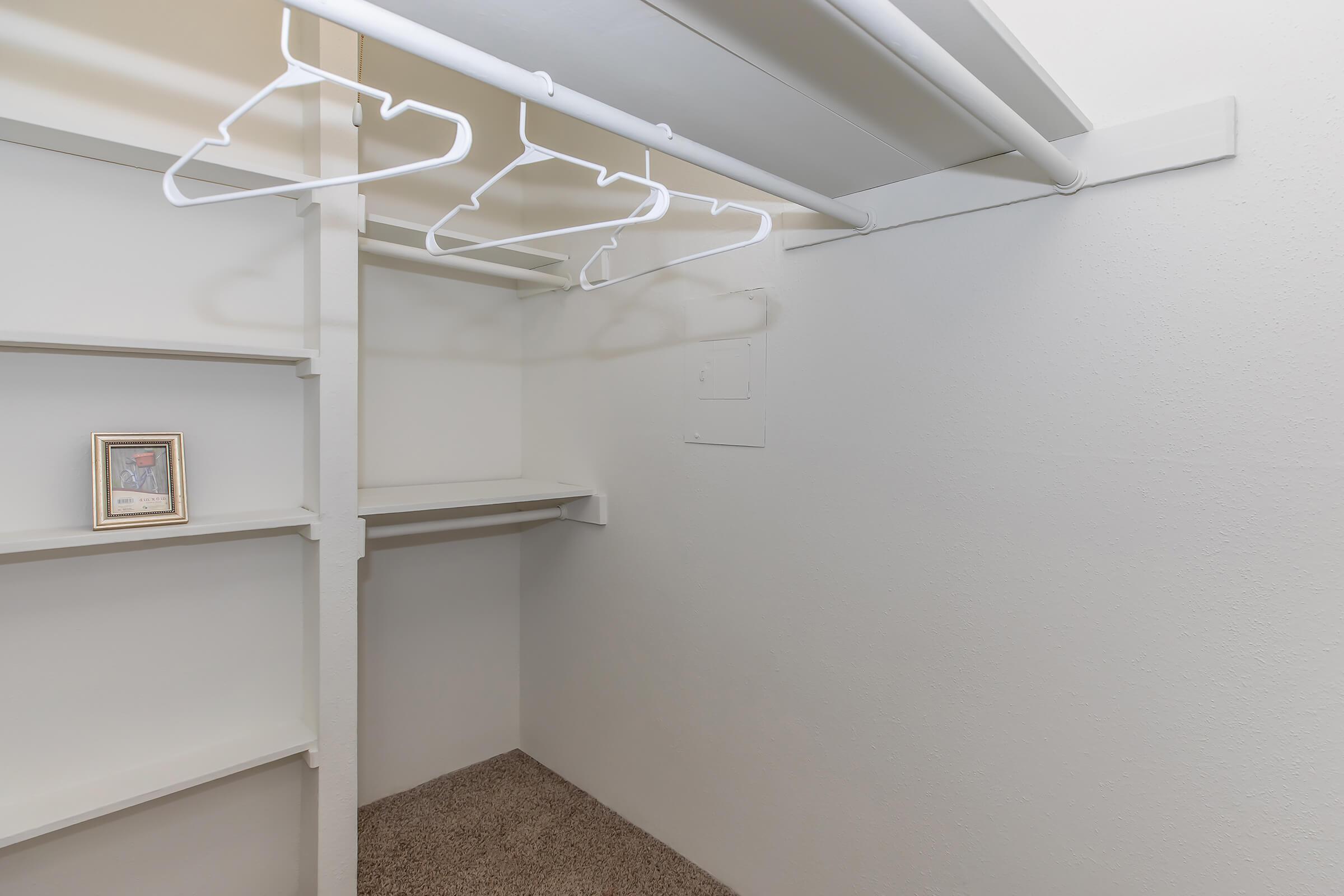 A clean, empty closet with white walls, featuring a long hanging rod with several empty white hangers. There are two shelves on the left side, and a framed photo sits on the bottom shelf. The floor is covered with a beige carpet.