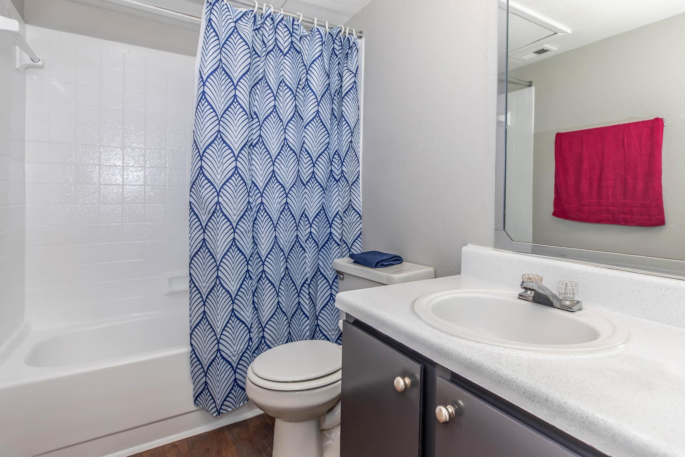 A bathroom featuring a white tub/shower combination, a blue and white leaf-patterned shower curtain, a white countertop sink, a toilet, a hand towel in red hanging on the wall, and a large mirror. The floor is a wood-like material, creating a warm atmosphere.