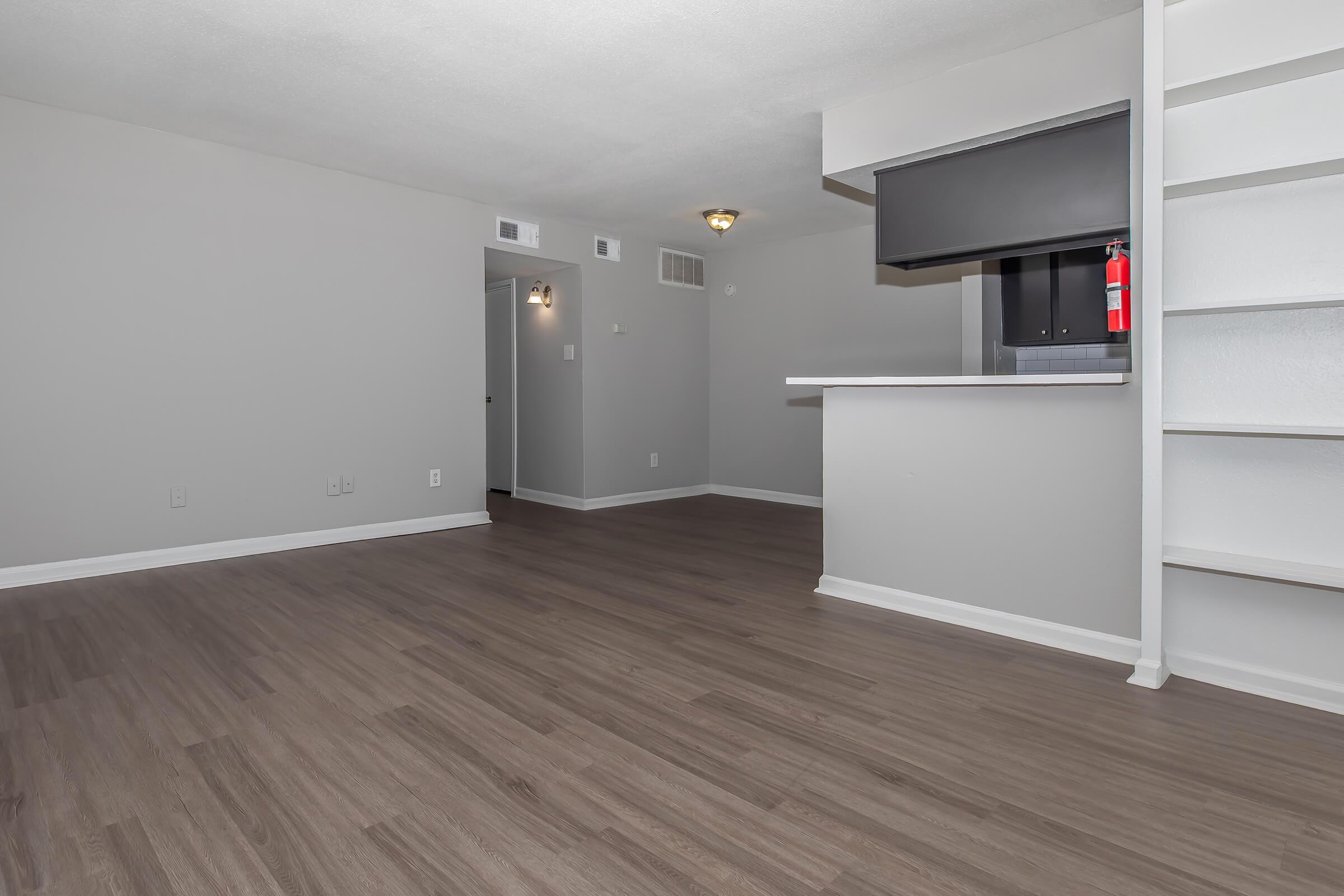 A spacious, well-lit room with gray walls and new wood-like flooring. There is a small kitchen area with a black countertop and an open shelf. The back wall features a door leading to another room. A fire extinguisher is mounted on the wall, indicating safety features. The overall ambiance is modern and inviting.