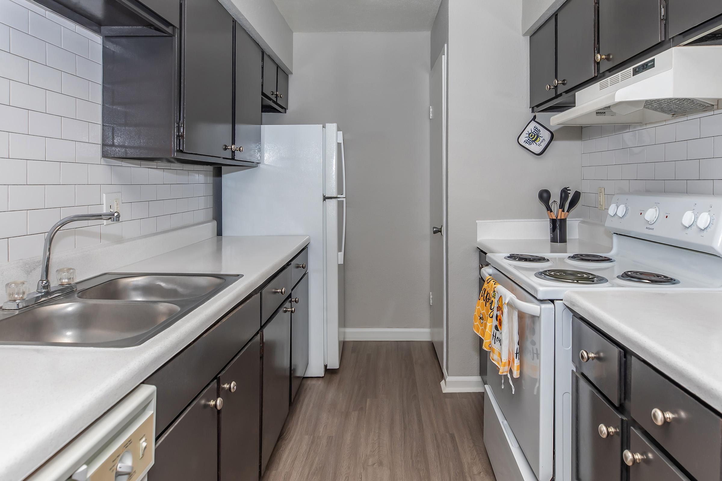 A modern kitchen featuring black cabinets, a white countertop, and stainless steel appliances. There's a double sink on the left, a stove and oven on the right, and a refrigerator in the background. The walls are light-colored, and utensils are neatly arranged near the stove.