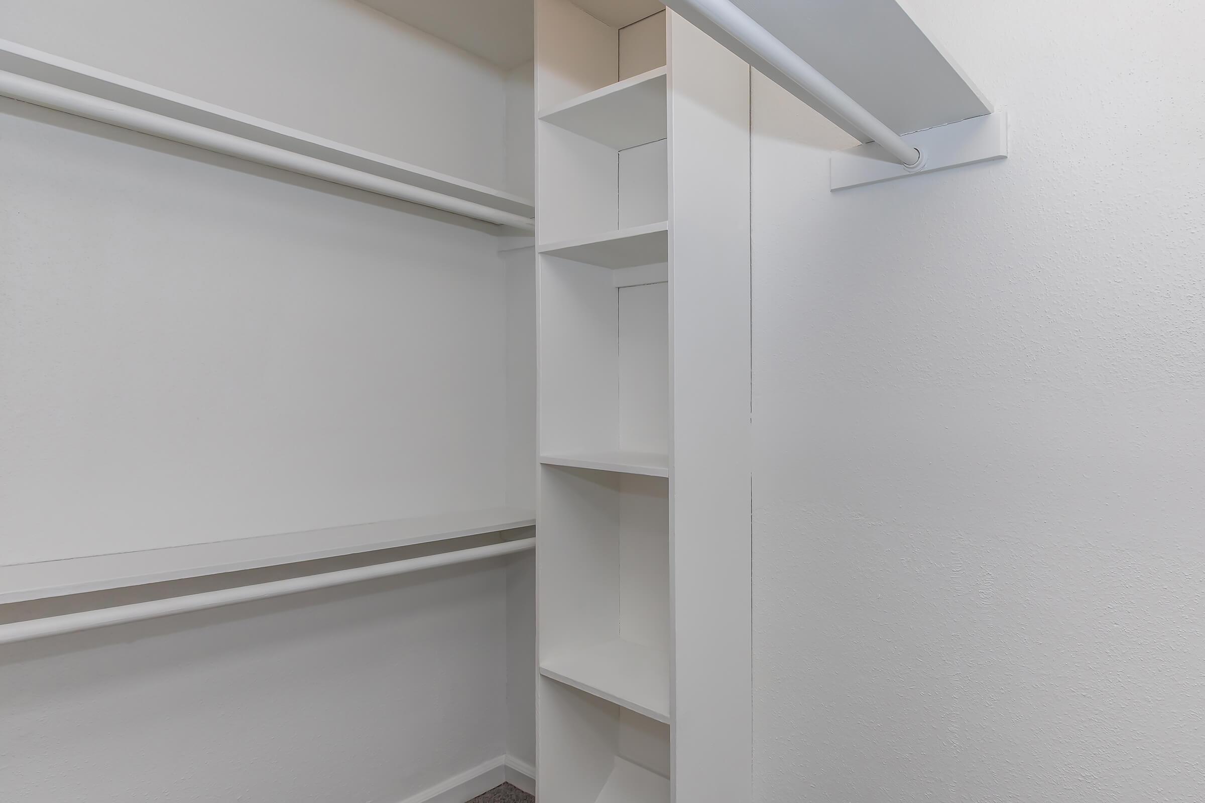 A clean and empty closet featuring white walls, a single shelf, and a vertical set of open shelves for storage. The space is well-lit and minimalistic, suggesting an organized and uncluttered environment.