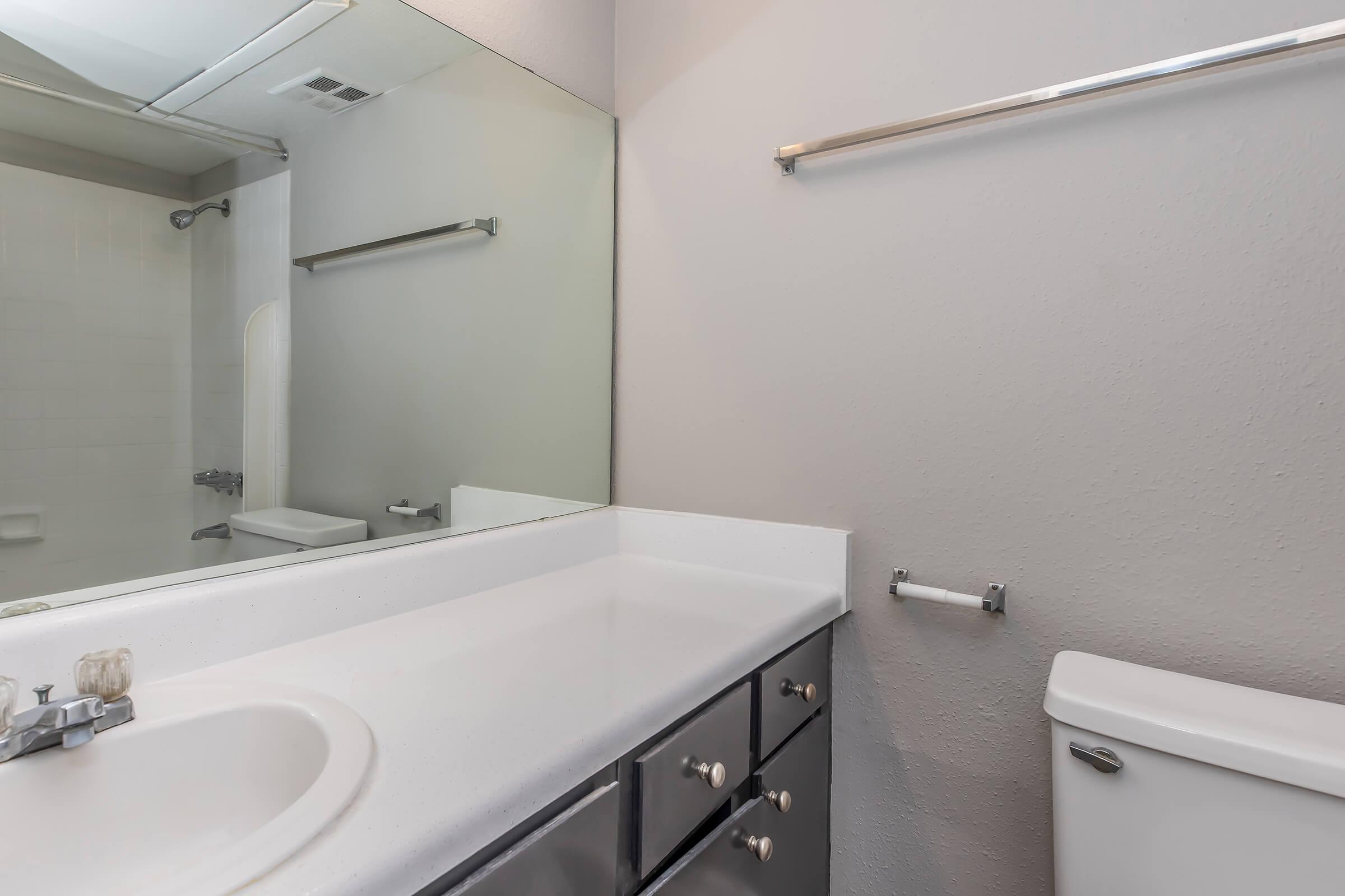 A clean and modern bathroom featuring a countertop sink with a mirror above, a toilet to the right, and a gray wall. A towel bar is mounted on the wall, and the space is well-lit and uncluttered. A glimpse of a shower area can be seen in the background.