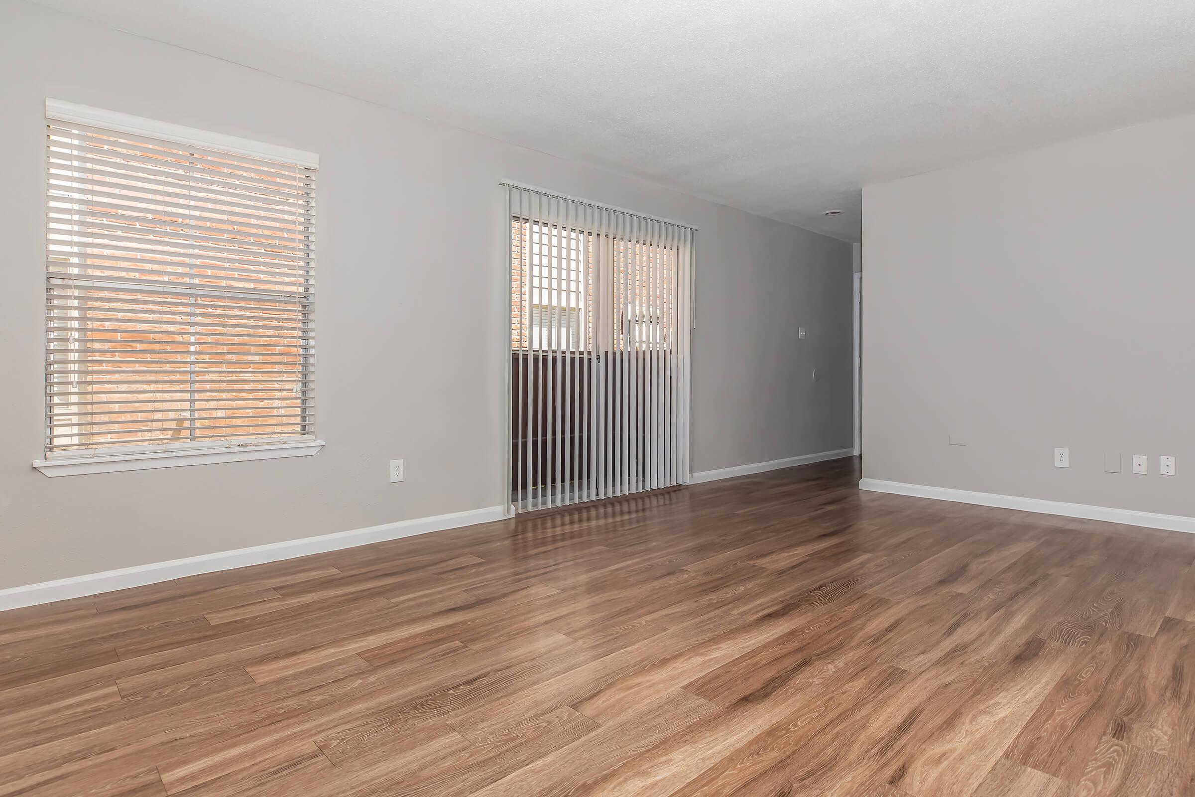 Interior of a spacious room featuring light-colored walls and hardwood flooring. There is a window with horizontal blinds on one wall, and another section with vertical blinds leading to a balcony. The room appears well-lit and empty, ready for furniture placement.