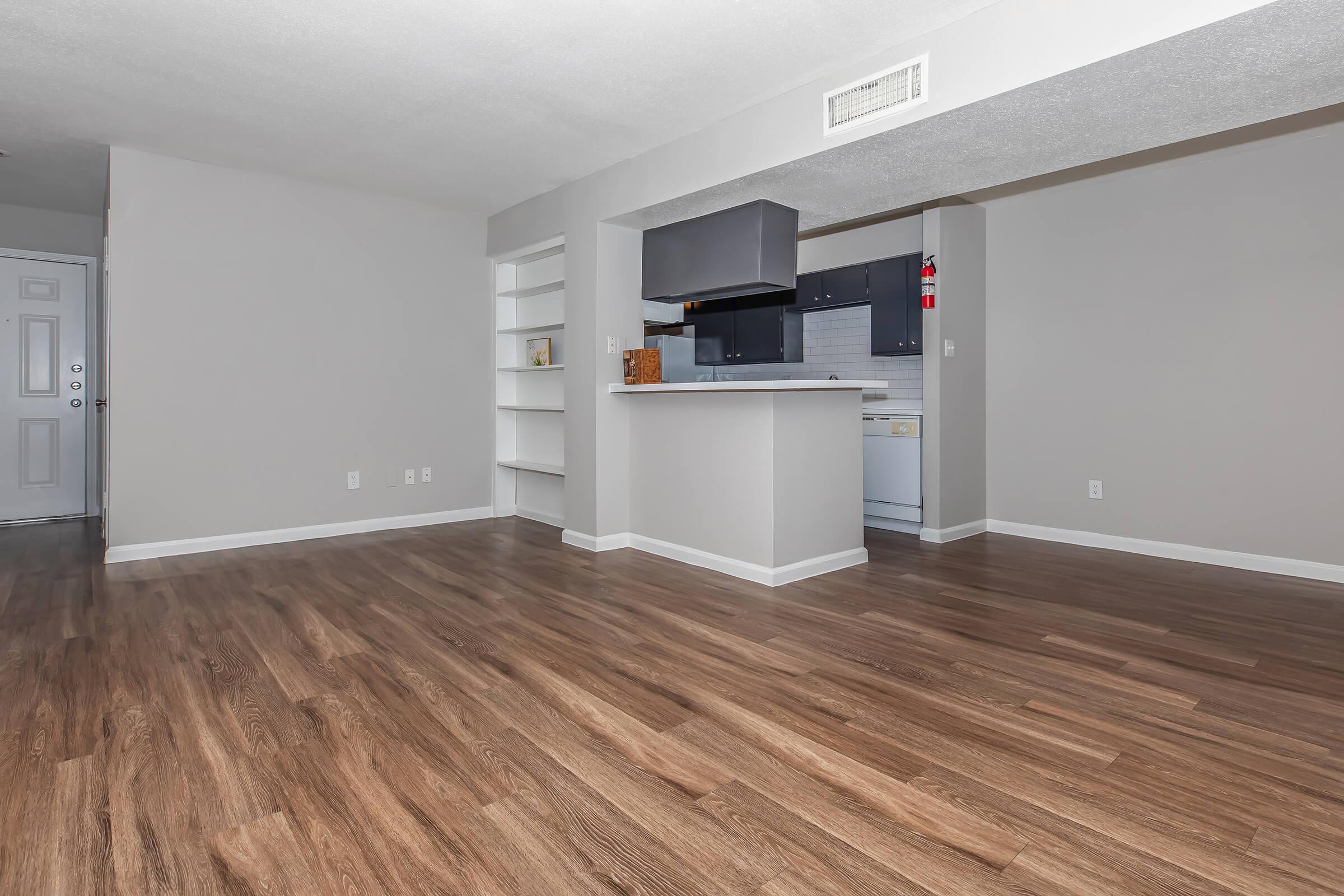 A spacious, empty living area featuring light-colored walls and modern laminate flooring. In the background, a small kitchen with dark cabinetry is visible, along with a white refrigerator and stove. A built-in shelf is situated on one side, and a front door is seen in the corner.