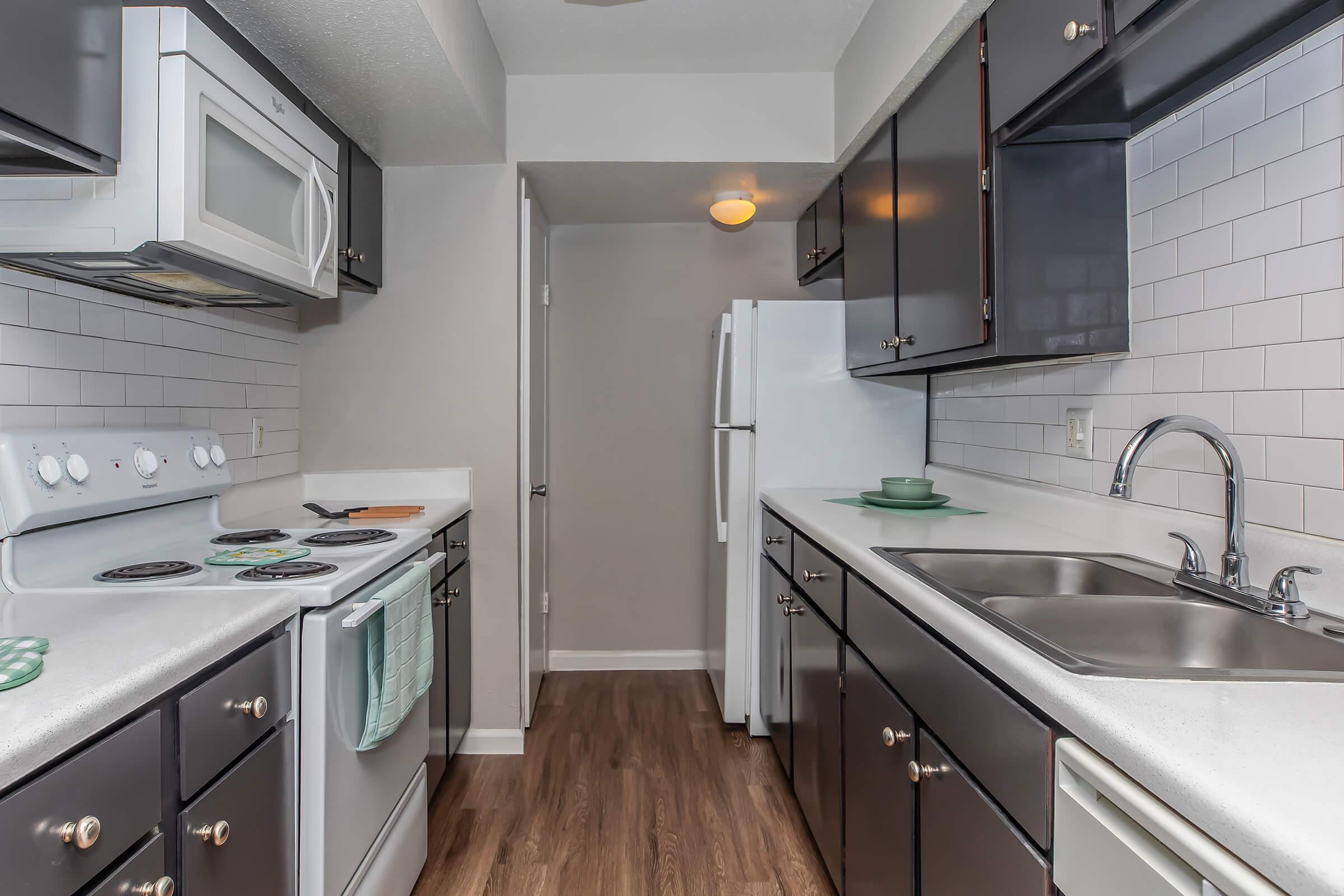 A modern kitchen featuring dark cabinetry, a white countertop, and stainless steel appliances. The kitchen includes a stove, oven, sink, and refrigerator, with a tiled backsplash and wood-style flooring, creating a clean and contemporary look.