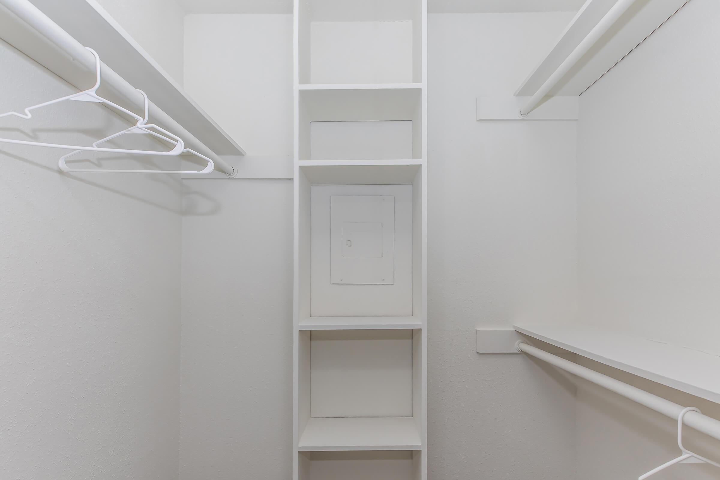 Empty closet with white walls, featuring built-in shelving and hanging rods. The design includes a tall vertical section with shelves and a utility panel in the center, providing organized storage space. Overall, the closet has a minimalist, clean appearance.