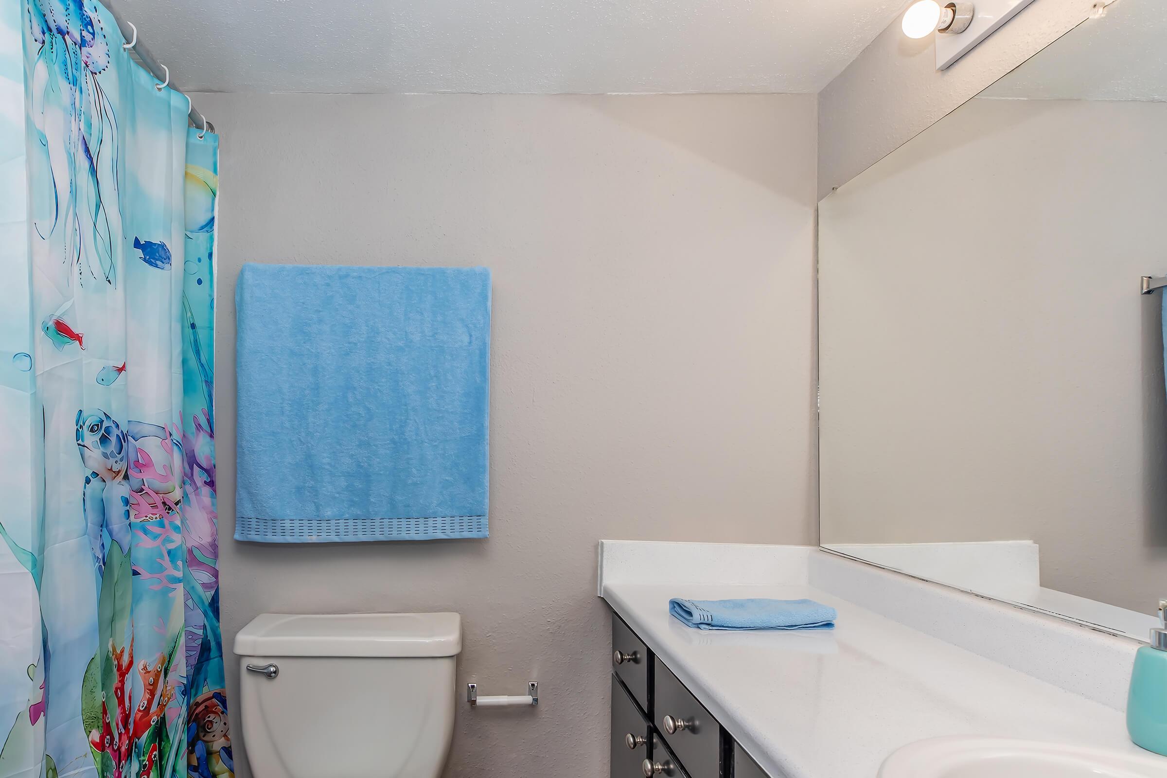 A clean bathroom featuring a white toilet, a large mirror above a sink with a light-colored countertop, and a colorful shower curtain decorated with marine life. Light blue towels hang on the wall, adding a pop of color to the neutral walls. A bathroom accessory is visible on the countertop.
