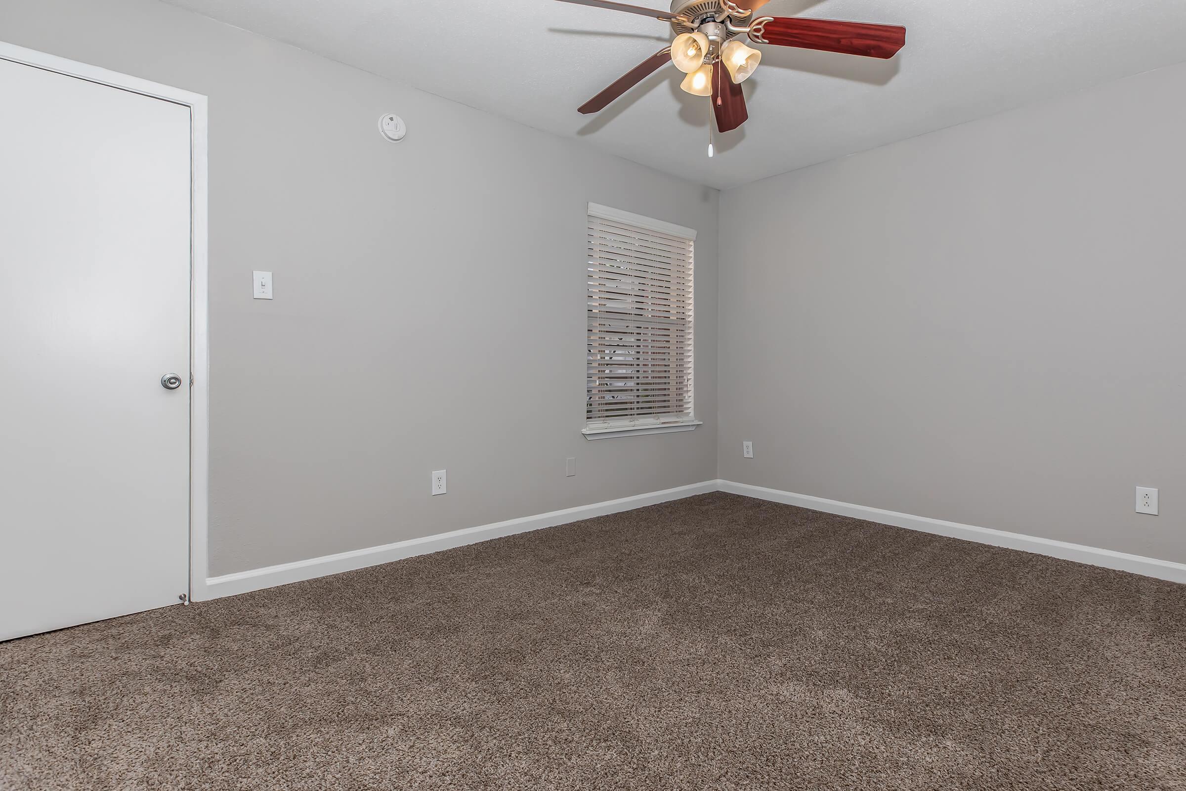 A light gray room with a ceiling fan and a large window with blinds. The floor is covered in light brown carpet. A white door is visible on the left, and the walls have no decorations, creating a clean and minimalist look.