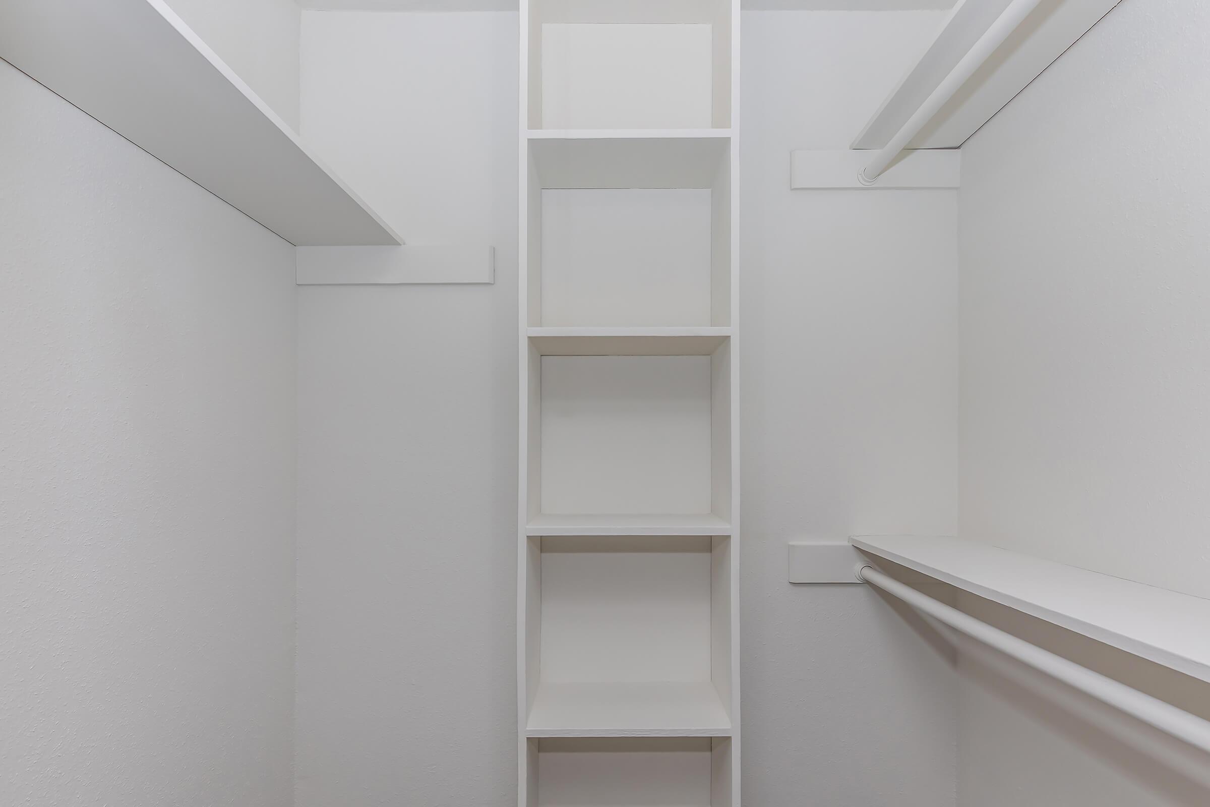 A minimalist closet with white walls and shelves. The closet features a tall, open shelving unit on one side and a hanging rod on the opposite side, providing space for organization and storage. The overall aesthetic is clean and modern.