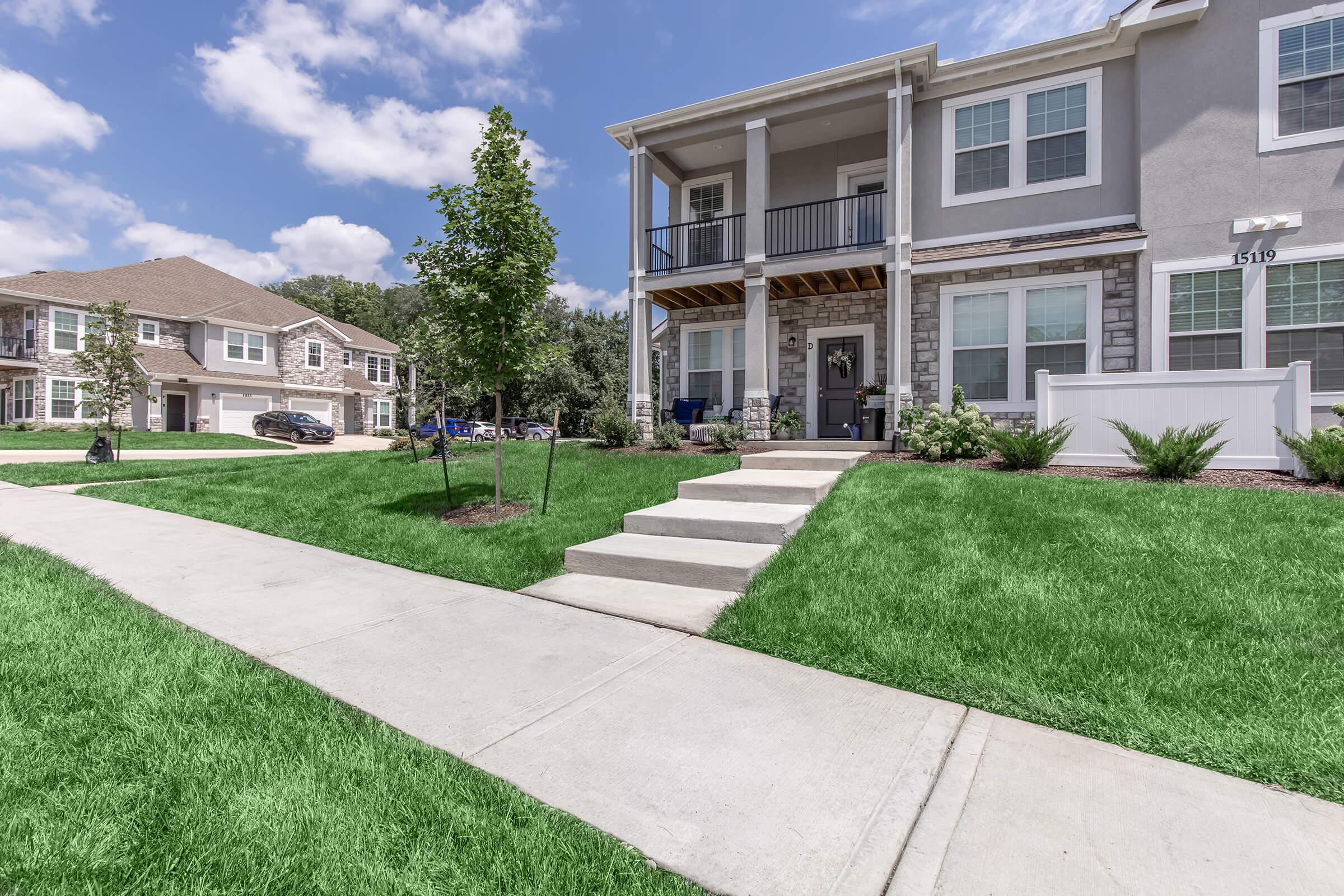 A well-maintained residential building with a covered porch and steps leading up to the entrance. The surrounding landscape features lush green grass, small trees, and a concrete sidewalk. In the background, additional residential units are visible under a partly cloudy blue sky.