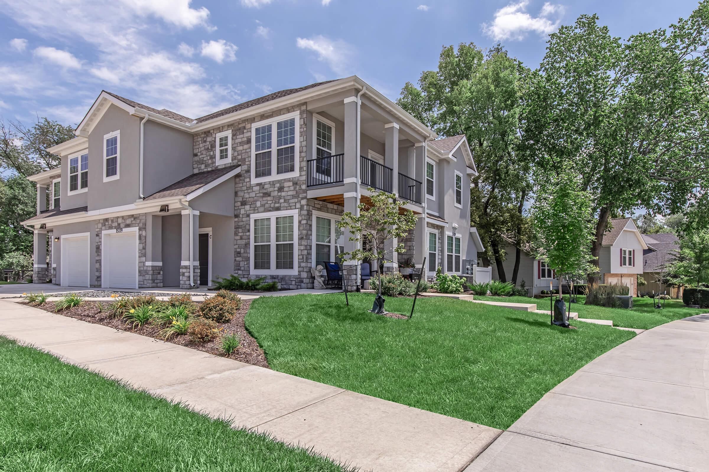 A modern two-story house with a stone and siding exterior. It features a front porch, large windows, and a well-maintained lawn with landscaping. A driveway leads up to the garage, and the surrounding area includes trees and shrubs under a blue sky with fluffy clouds.