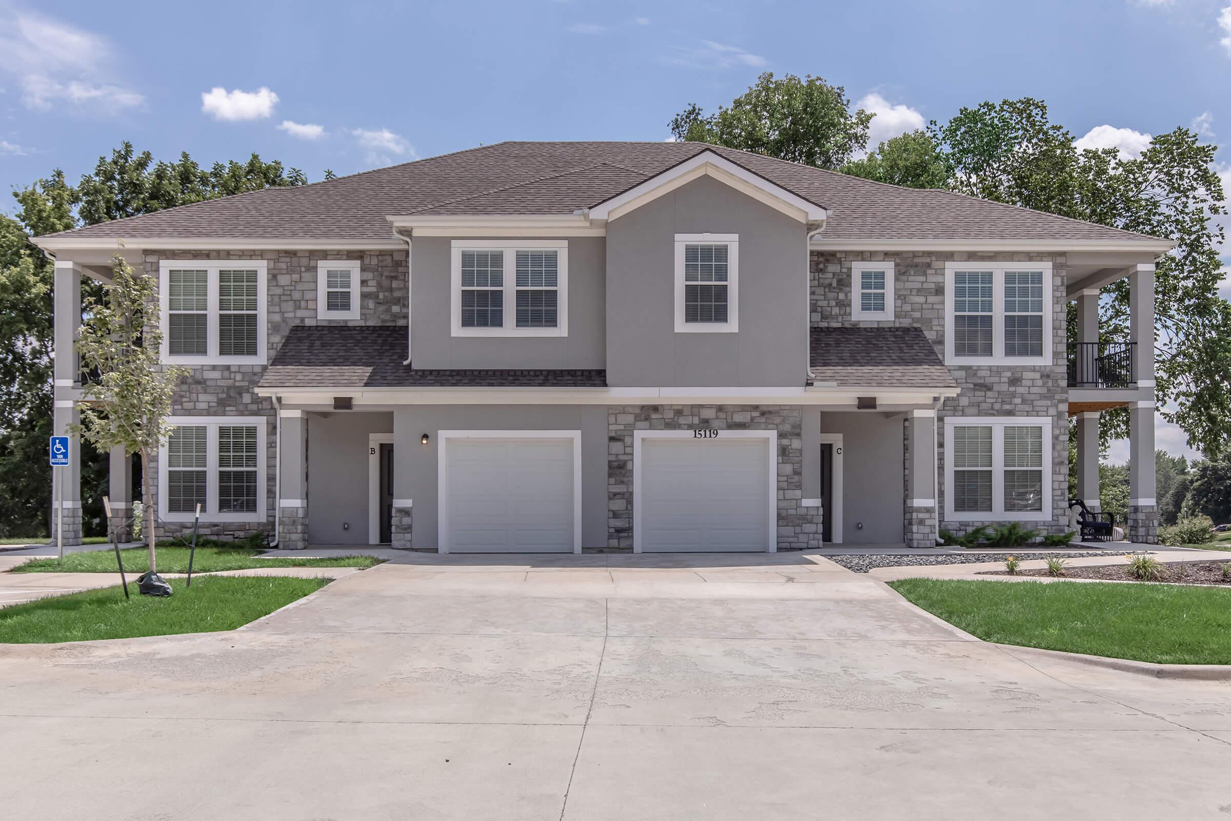 A two-story residential building with a stone and gray exterior. It features a central entrance, multiple windows, and two garages. The surrounding area has green grass, trees, and a clear blue sky. The driveway is paved and leads to the garages on the ground level.