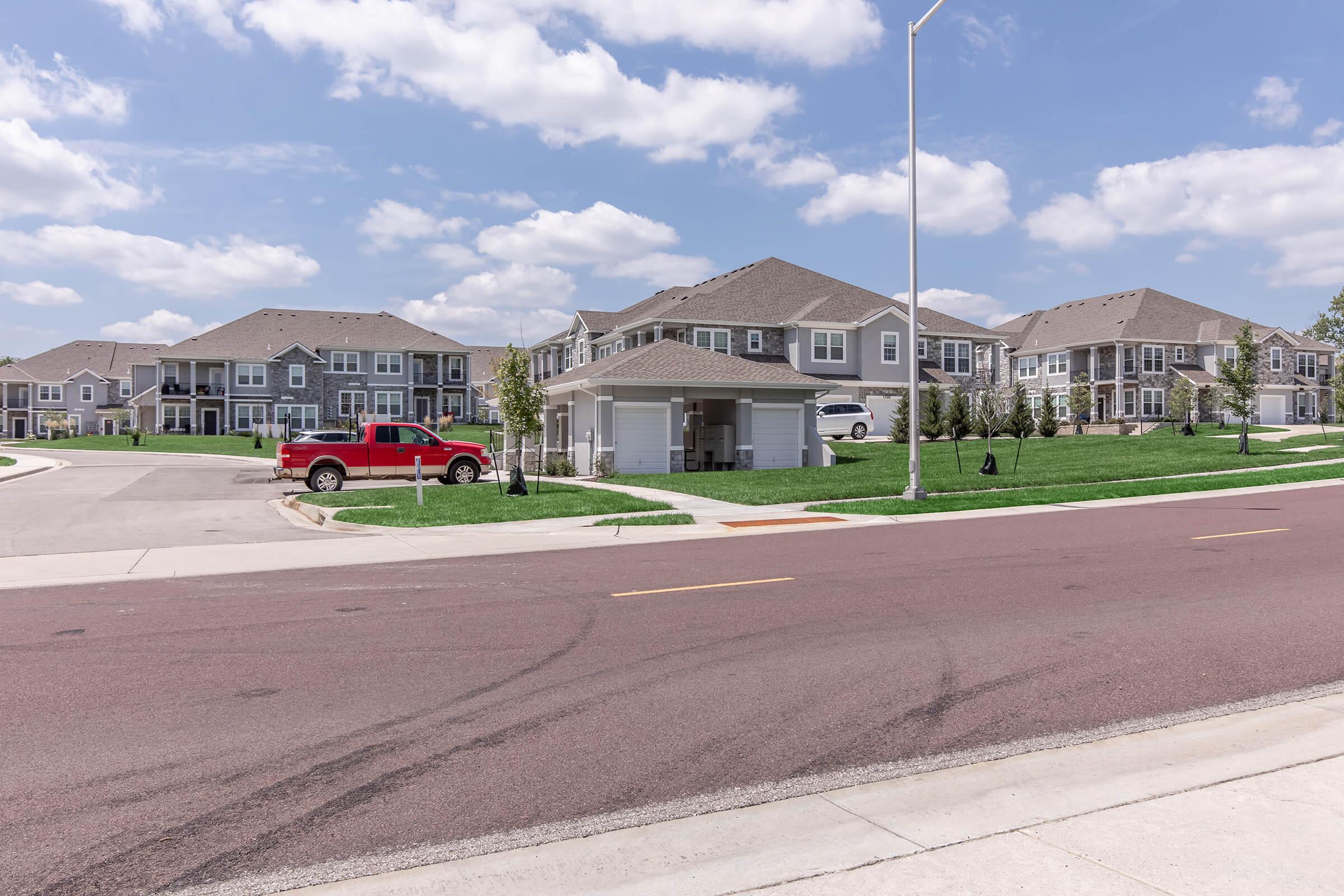 A residential neighborhood featuring several modern houses, a red truck parked in front of a garage, and well-maintained green lawns. The sky is partly cloudy, and the layout includes a curved street leading into the area.