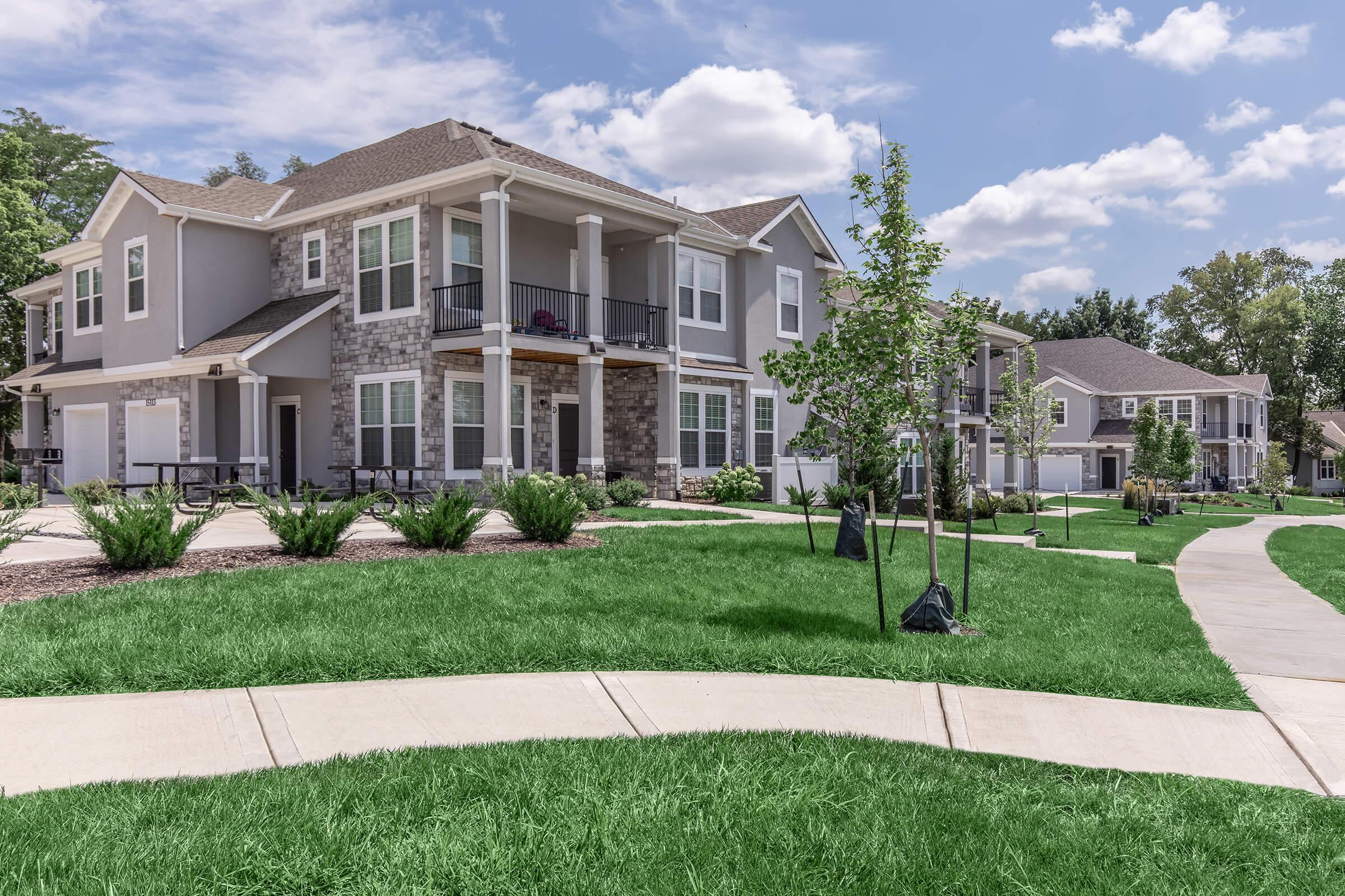 A residential area featuring modern apartment buildings with stone accents and balconies. The landscape is well-kept with green grass, neatly trimmed shrubs, and walking paths curving through the scene. In the background, there are trees and a partly cloudy sky, creating a serene atmosphere.