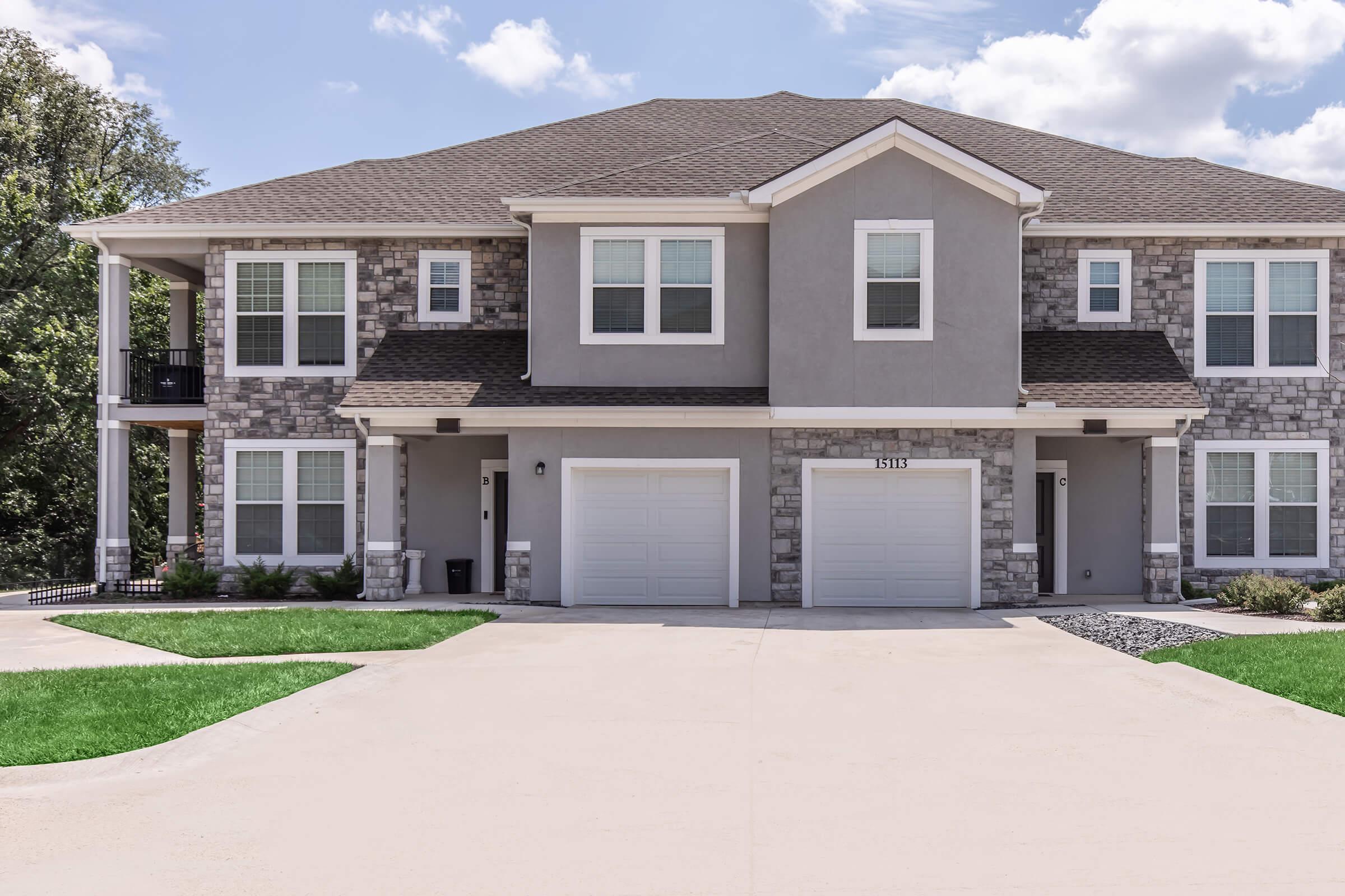 A modern two-story duplex with a stone and gray exterior. The building features two garages, multiple windows, and a landscaped front yard. The driveway is paved, and the surrounding area includes green trees and blue skies with fluffy white clouds.