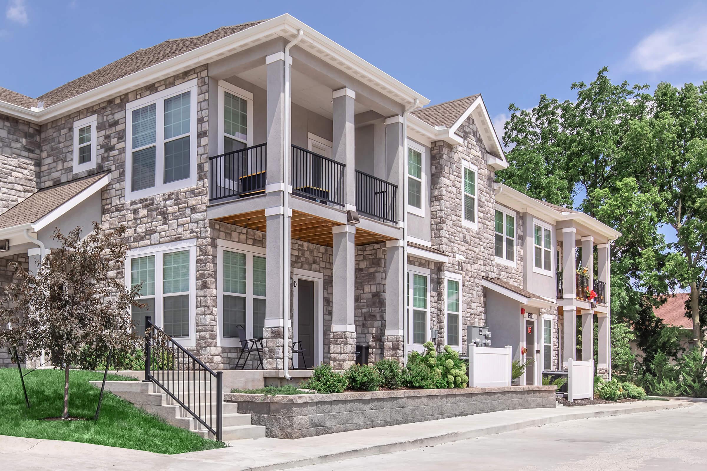 A modern multi-story residential building featuring stone accents, covered porches with railings, large windows, and well-maintained landscaping. The setting appears bright and inviting, with a clean path leading to the entrance and greenery around the property.