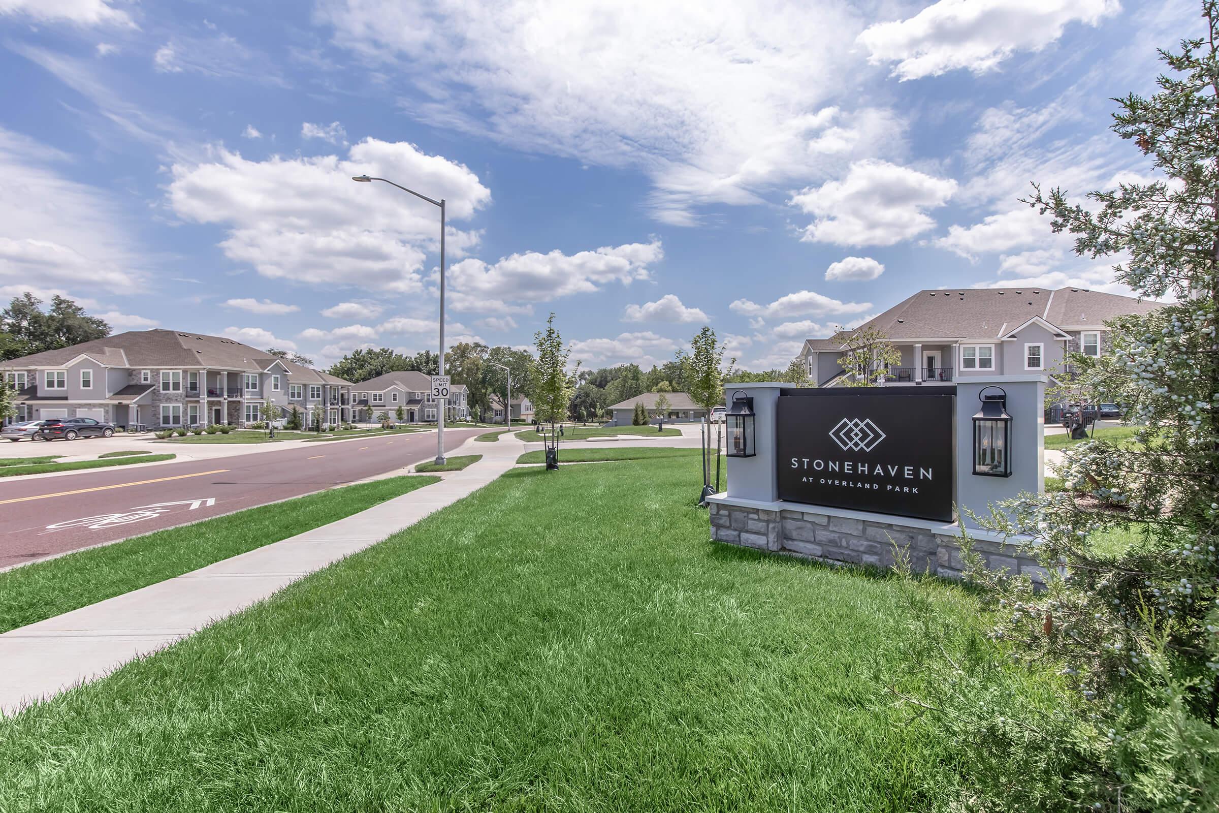 A suburban community entrance sign for Stonehaven at Windermere Park is displayed. The sign is surrounded by well-manicured grass, with a tree nearby. In the background, there are residential buildings and a clear blue sky with scattered clouds. The scene conveys a peaceful neighborhood atmosphere.