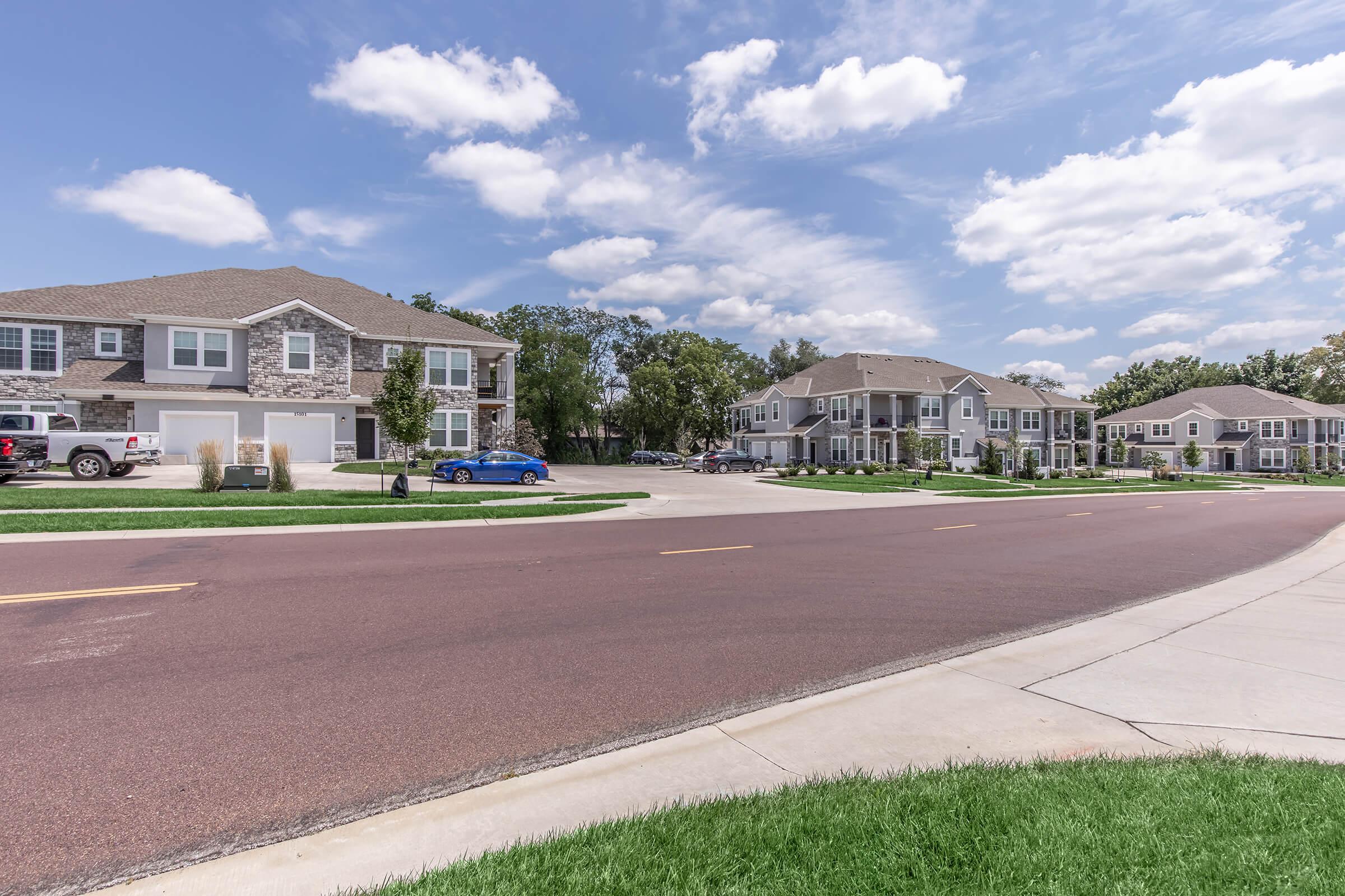 A quiet residential street lined with modern townhouses, featuring stone and wood exteriors. The road is newly paved and curves gently, with green grassy patches and a few parked cars visible. Fluffy white clouds float against a bright blue sky, creating a serene suburban atmosphere.