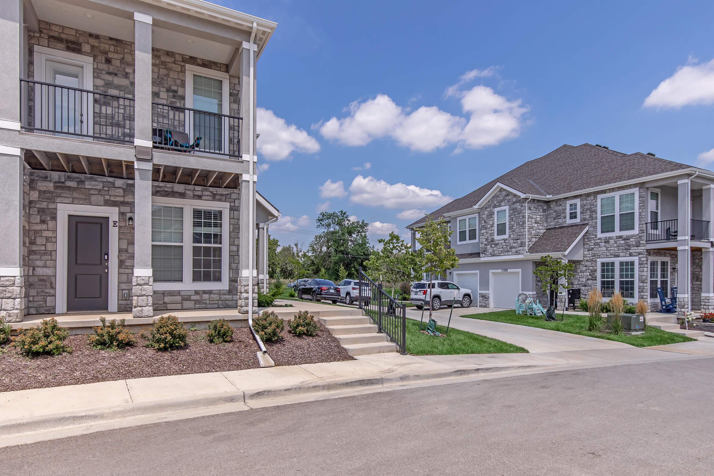 A scenic view of a suburban neighborhood featuring two stone-faced houses with balconies, well-maintained lawns, and a clear blue sky. The foreground includes driveways with parked cars, and the overall atmosphere is calm and inviting, showcasing a blend of residential architecture and greenery.