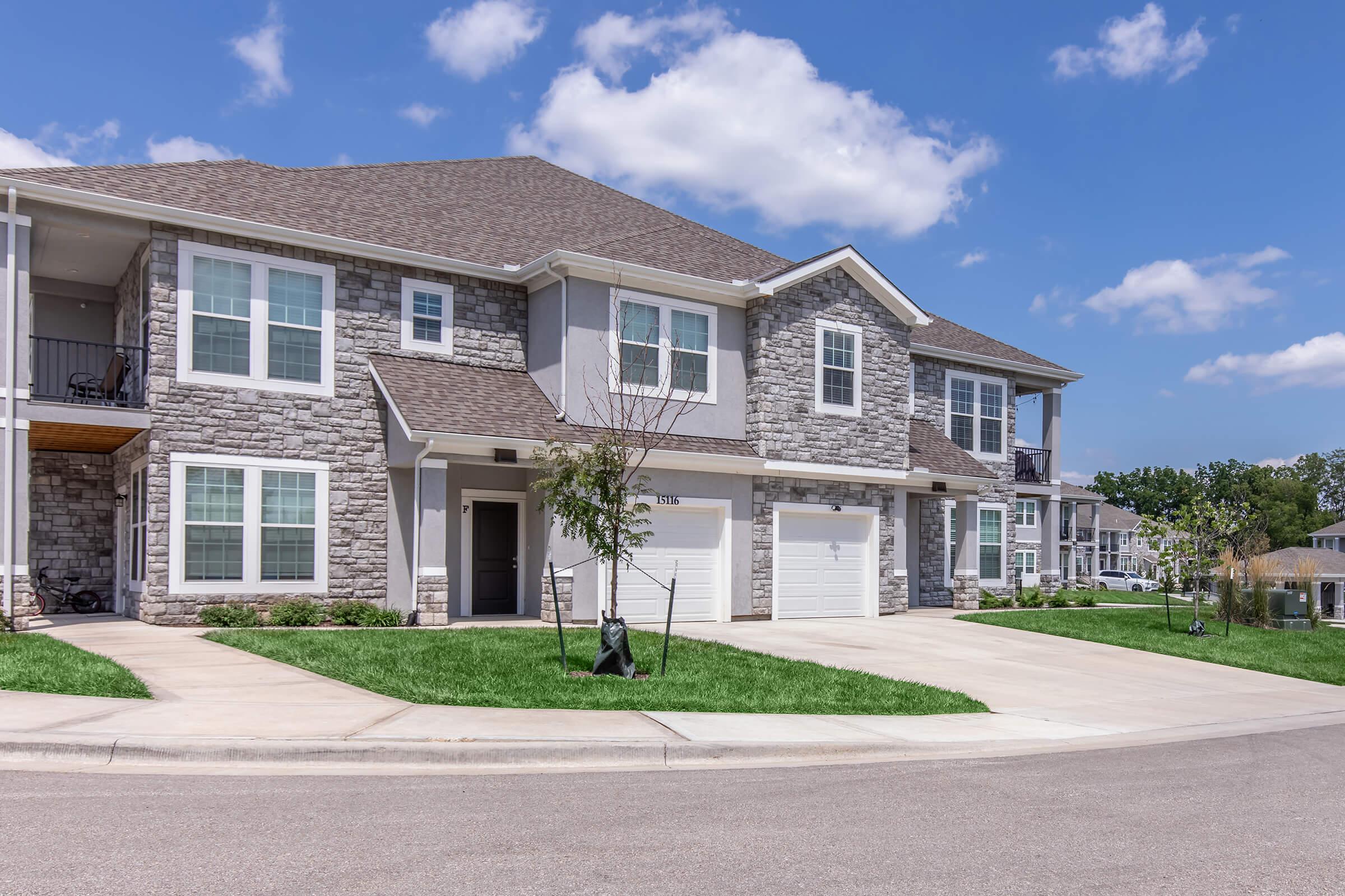 A modern, two-story residential building with stone and siding exterior. The property features several large windows and a driveway leading to two garages. Green grass surrounds the building, and a few trees are planted in the area. The sky is bright with scattered clouds.
