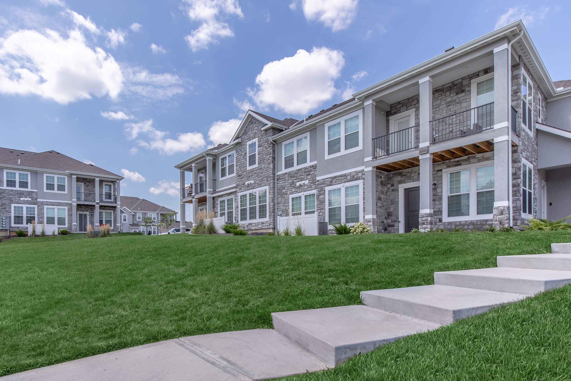 A modern apartment complex featuring two multi-story buildings with stone accents and large windows. The foreground has a well-maintained green lawn and concrete walkways leading to the entrances of the apartments, under a partly cloudy blue sky.