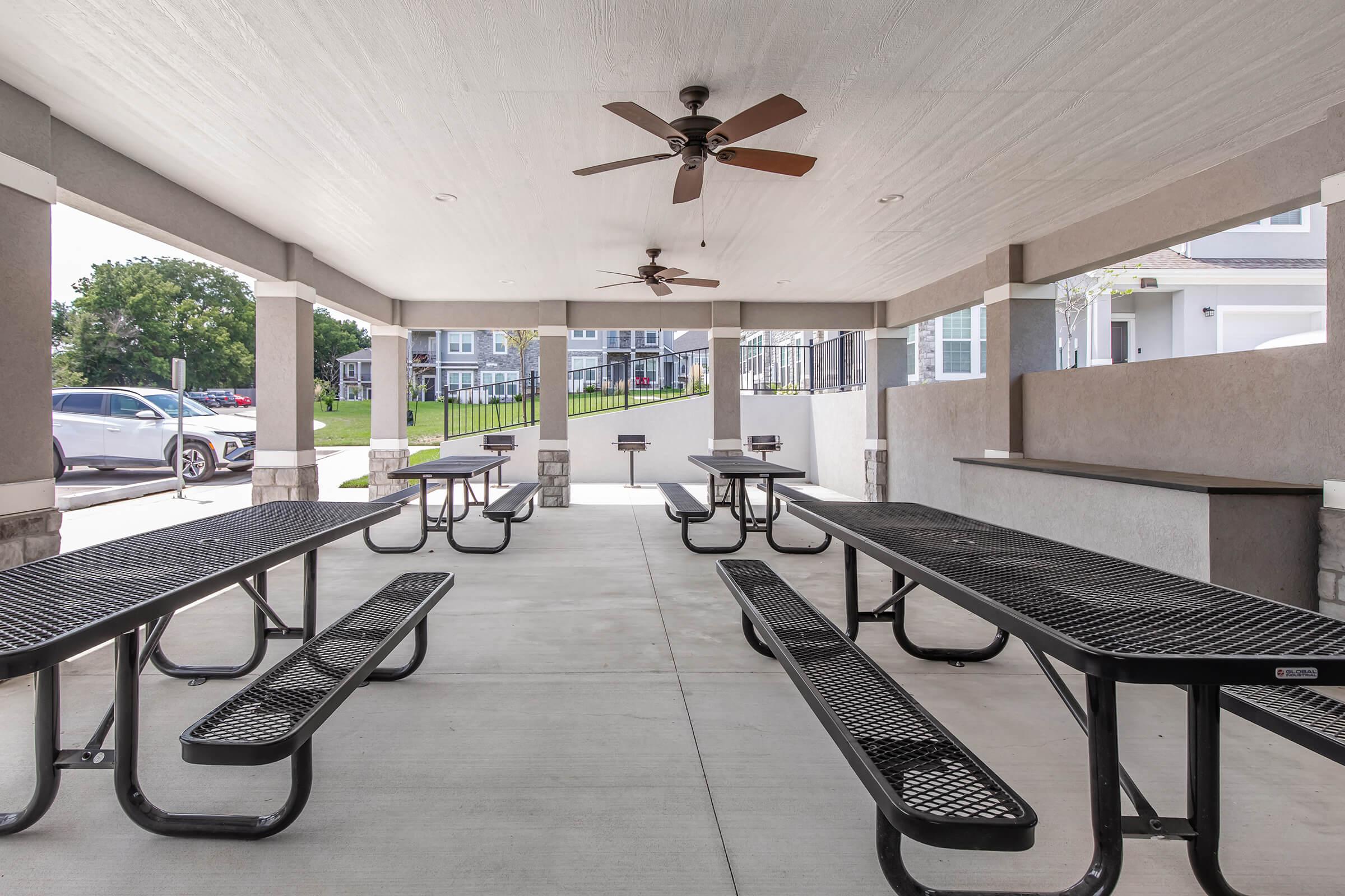 Covered outdoor seating area with multiple black picnic tables and fans mounted on the ceiling, situated in a well-lit space. The background features a grassy area and a building structure visible through the open sides.