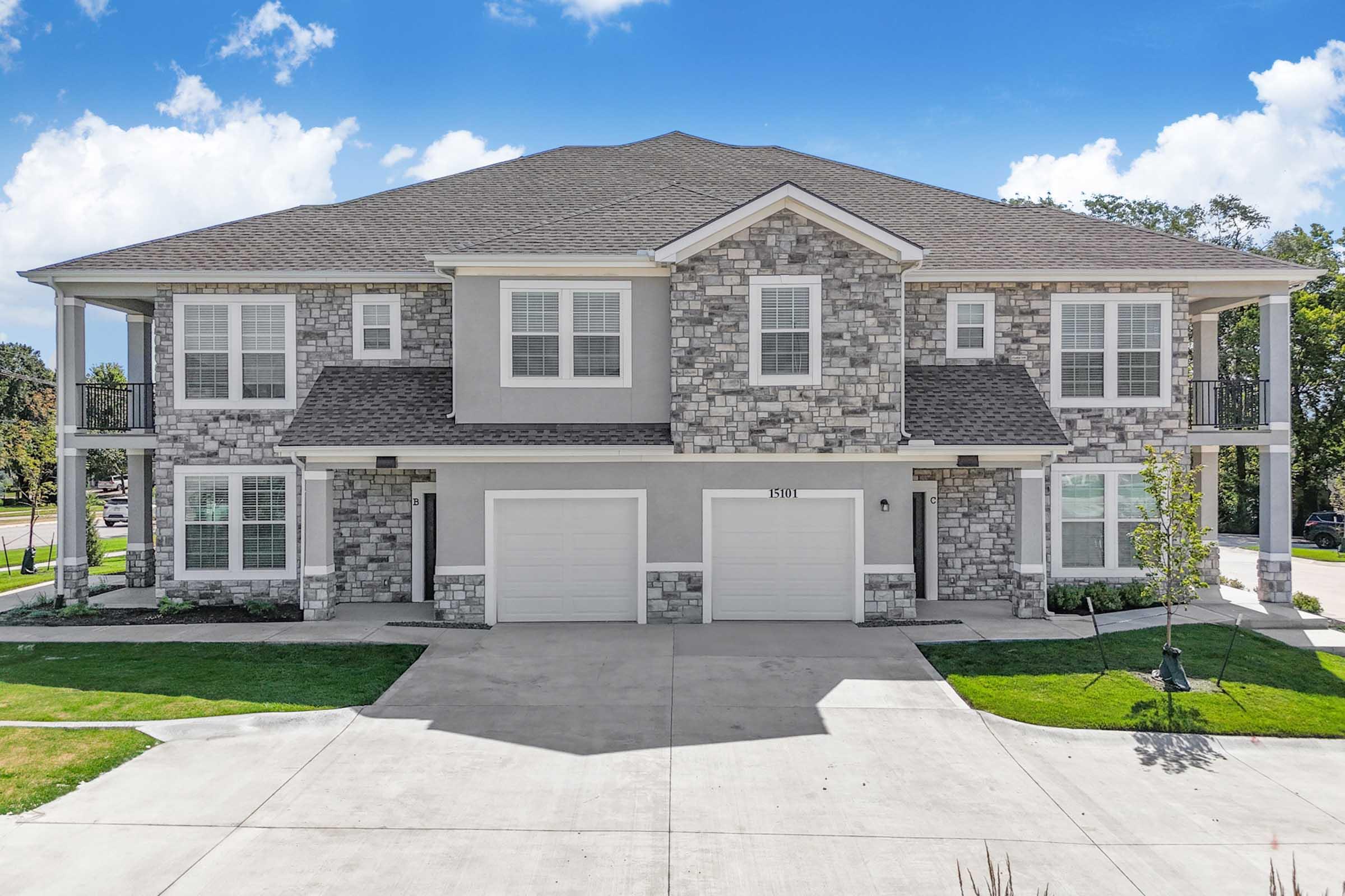 A modern two-story apartment building featuring a stone facade, large windows, and front garages. The structure has a well-maintained lawn with green grass and small trees. The sky is clear with a few clouds, creating a bright and inviting atmosphere.