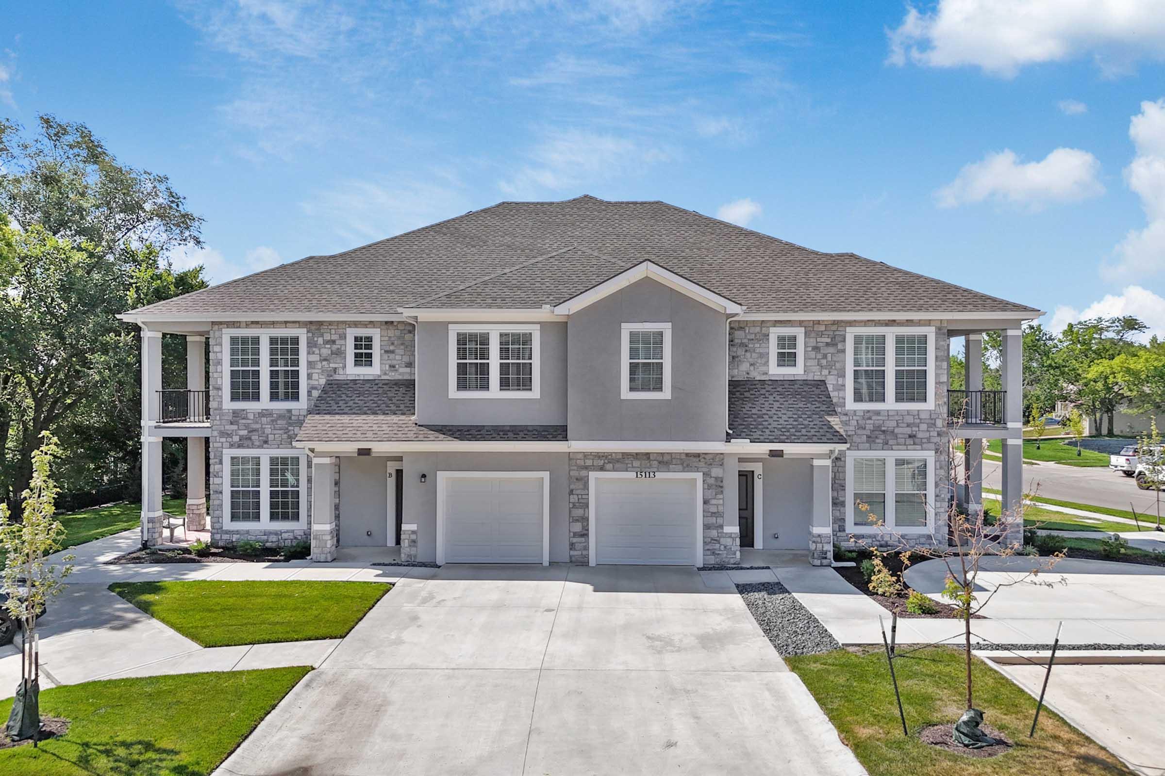 A modern two-story duplex featuring a stone and siding exterior. The building includes multiple windows, a front porch with railings, and two garages. The well-maintained lawn has trees and landscaping, set against a clear blue sky.